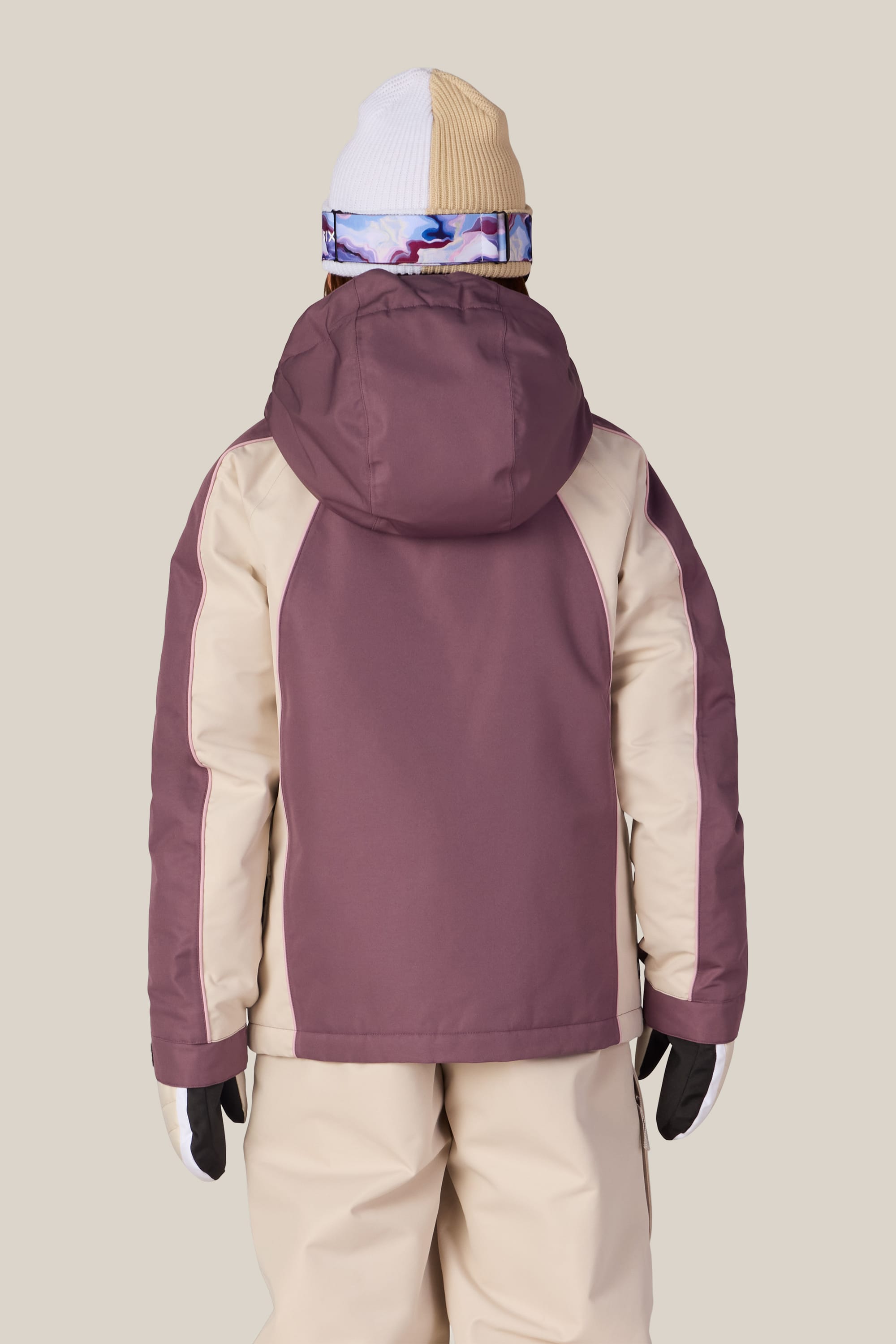 A person is wearing the 686 Girls’ Hologram™ Insulated Jacket by 686 in a two-tone mauve and beige colorway, paired with beige pants, gloves, a white and beige knit hat, and a colorful neck warmer. They are facing away from the camera against a plain background.
