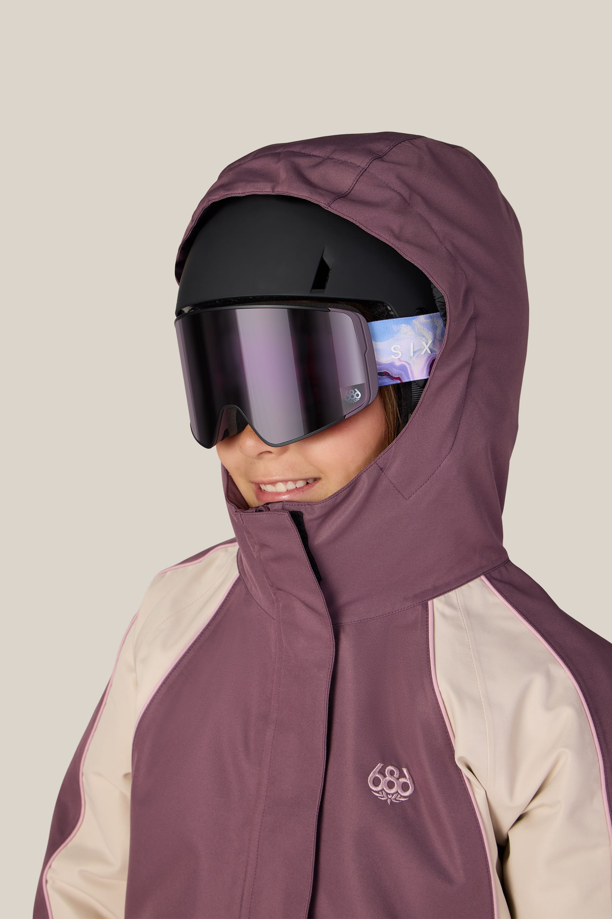 A child wearing the 686 Girls’ Hologram™ Insulated Jacket by 686, featuring a mauve and cream woven design with a hood, along with a black ski helmet and reflective ski goggles, smiles slightly against a plain background.