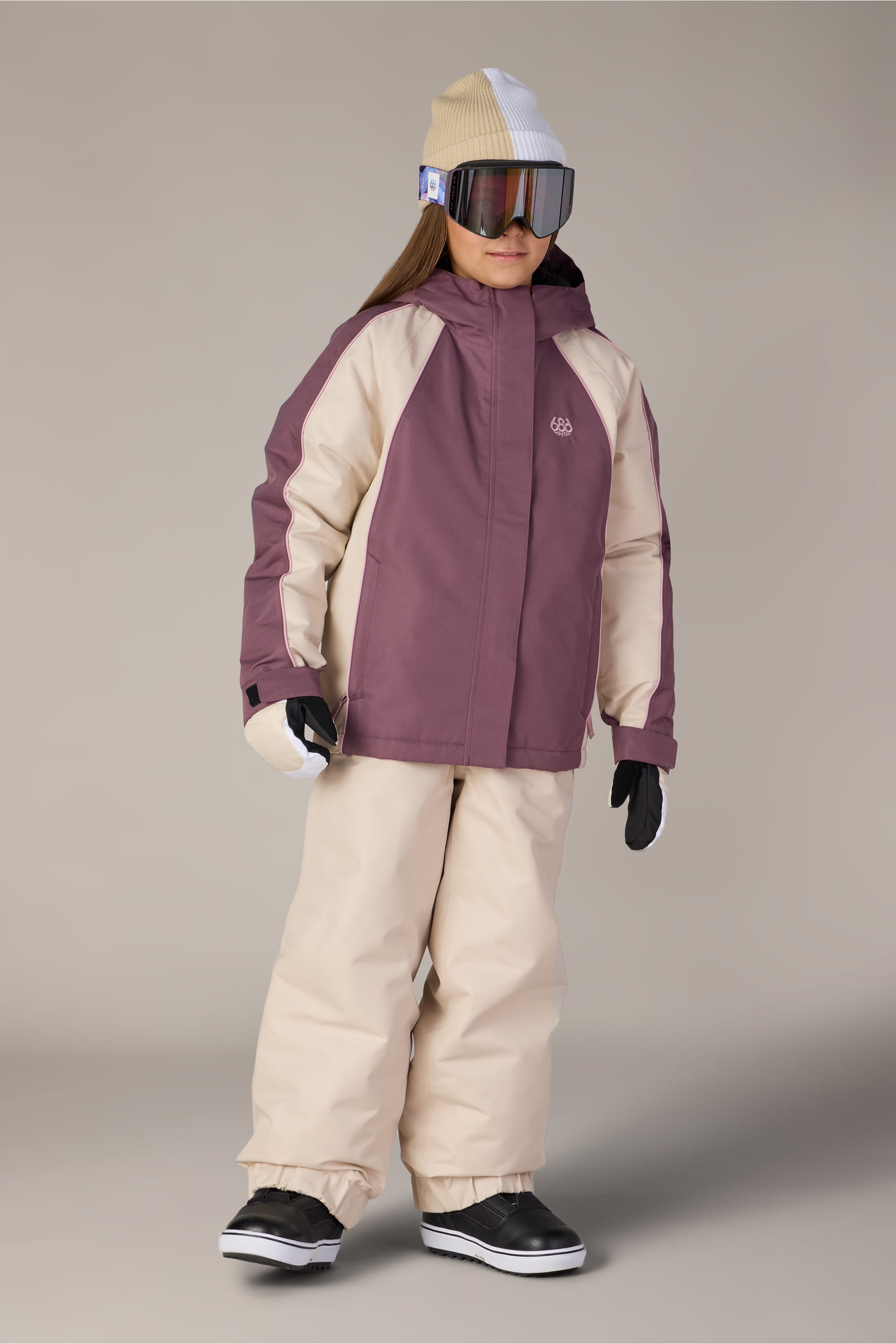 A child wearing the 686 Girls’ Hologram™ Insulated Jacket in purple and beige, paired with matching waterproof pants, a beige and white beanie, large ski goggles, black gloves, and black snow boots, standing against a plain background.