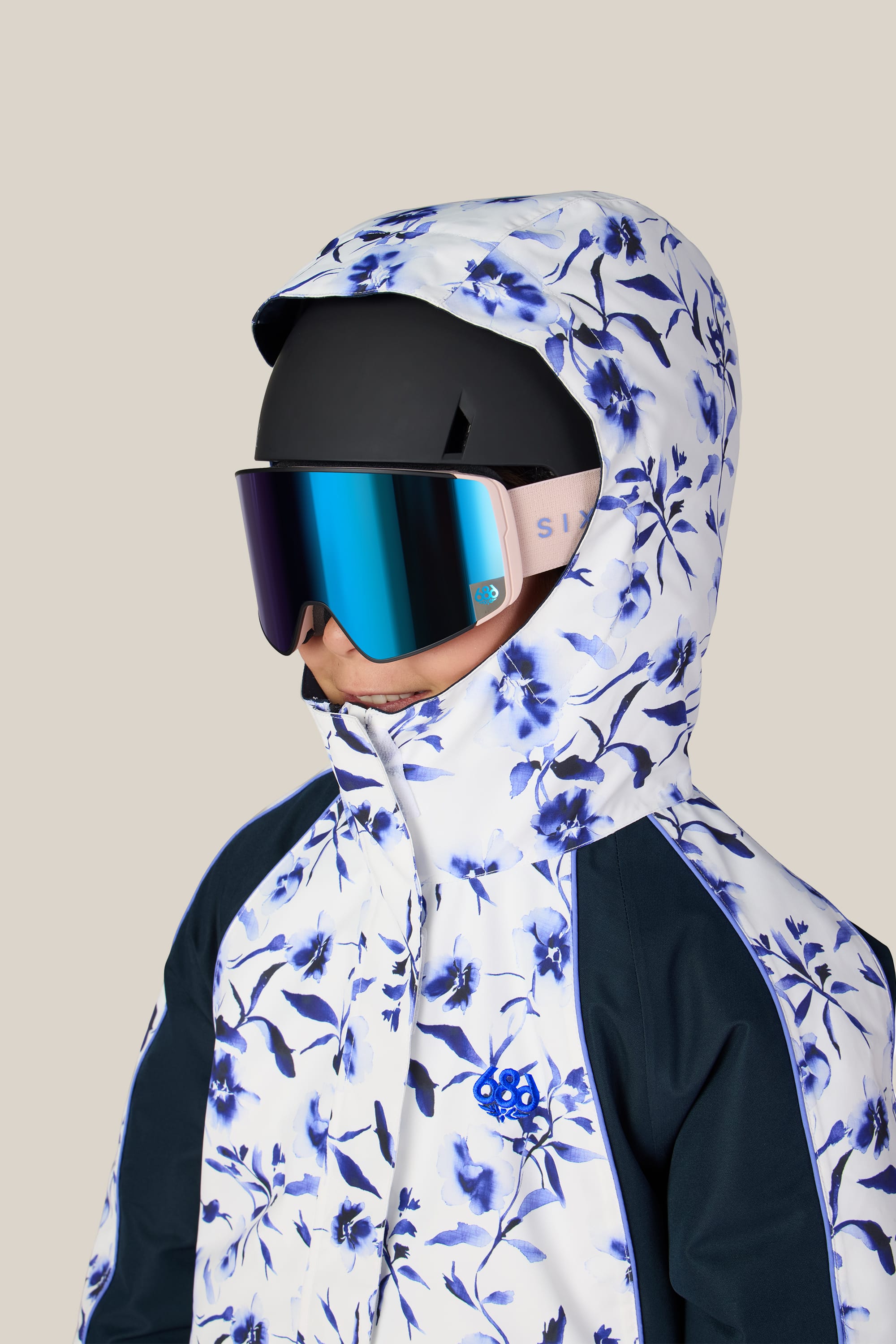 A person is wearing the 686 Girls’ Hologram™ Insulated Jacket by 686, featuring a blue floral pattern and hood, paired with a black ski helmet and reflective ski goggles against a plain beige background.