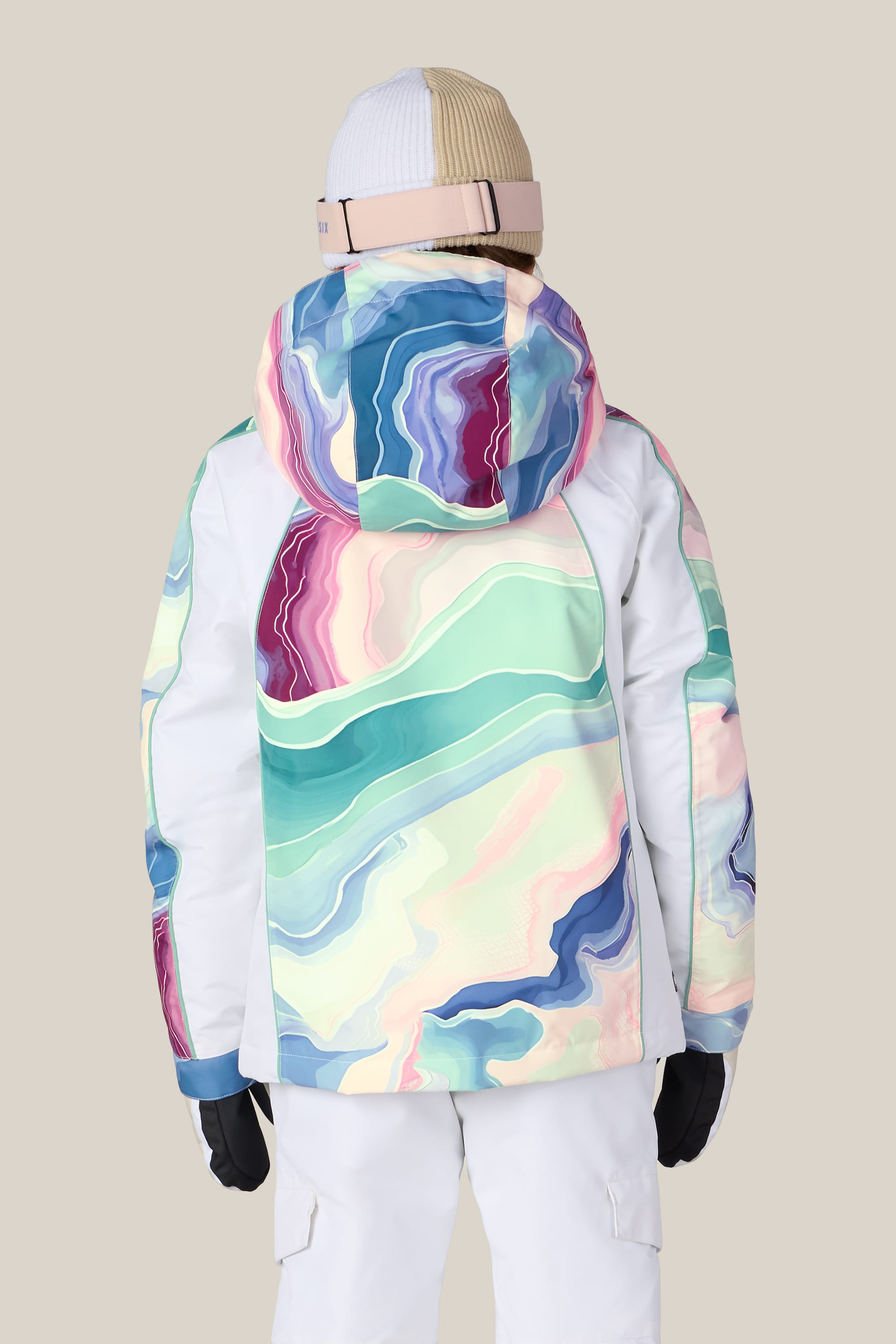 A girl is seen from behind wearing the 686 Girls’ Hologram™ Insulated Jacket by 686, featuring a colorful abstract pattern, paired with white pants, a beige and white beanie, and ski goggles, standing against a plain light background.