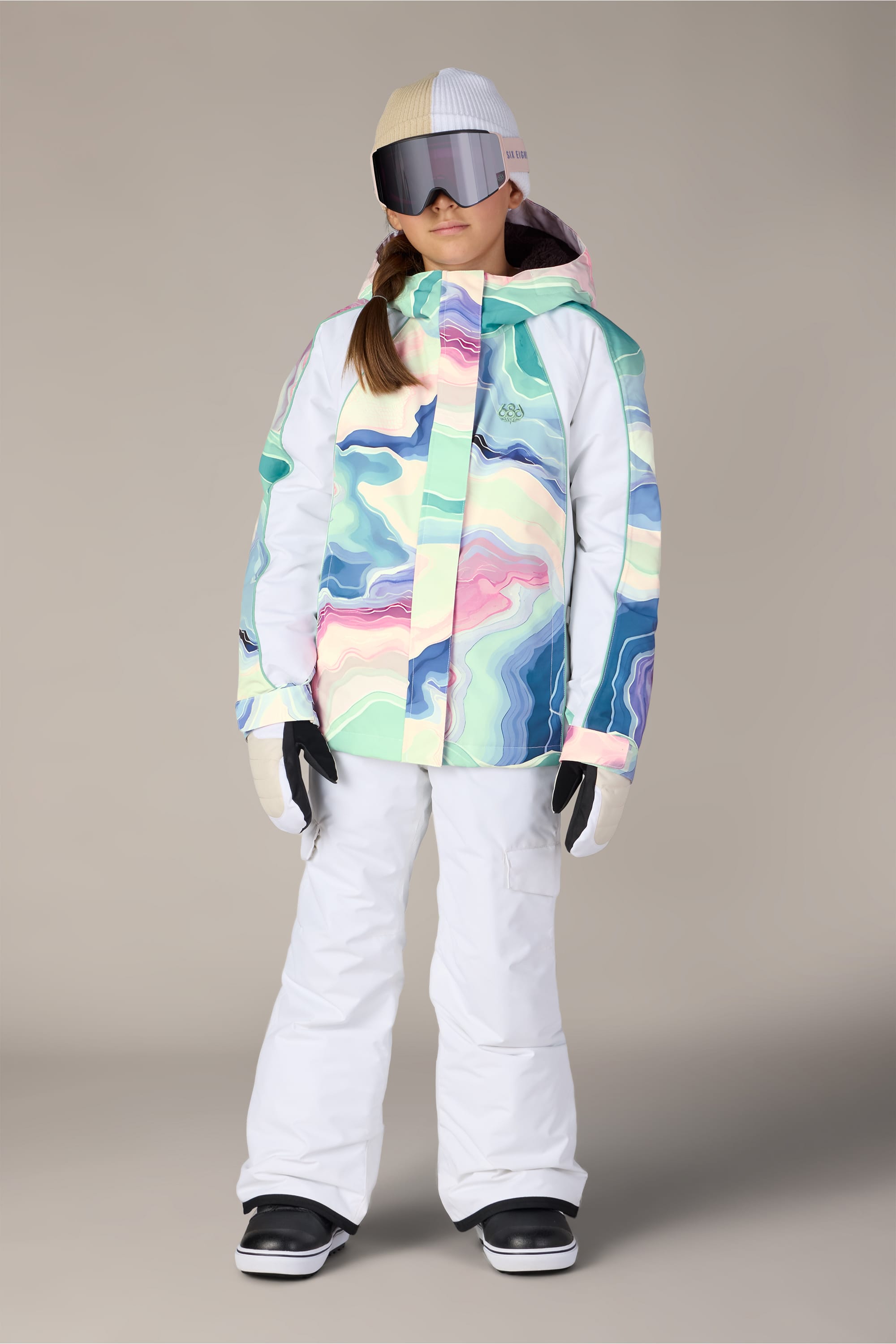 Wearing the 686 Girls’ Hologram™ Insulated Jacket—a colorful pastel-patterned waterproof design from 686—a person stands against a plain background, paired with white snow pants, gloves, black boots, large reflective ski goggles, and a white beanie.