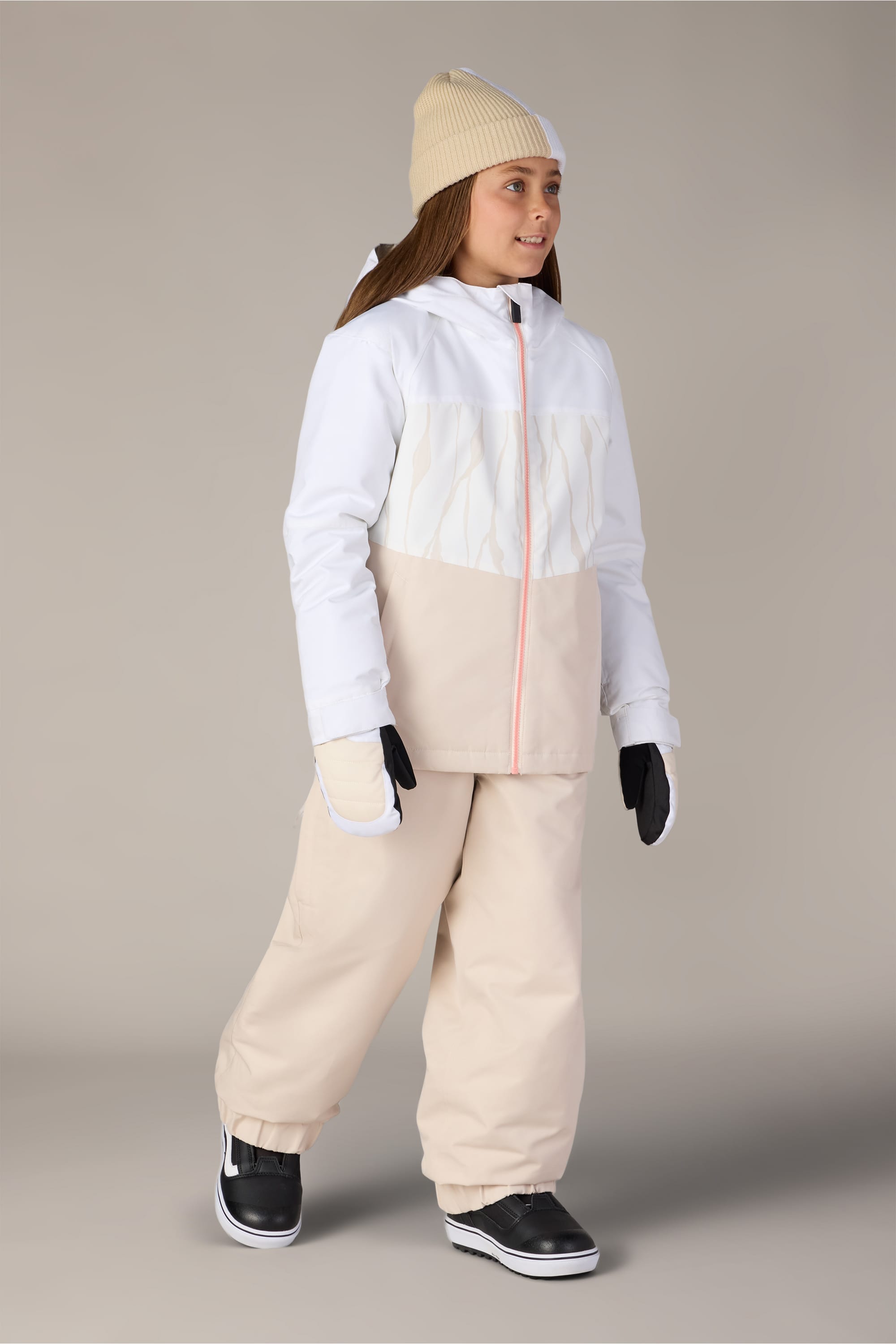 A girl wearing the 686 Girls’ Outline™ Insulated Pant in beige, paired with a white and beige winter jacket, a beige beanie, black gloves, and black boots stands and smiles against a plain gray background.