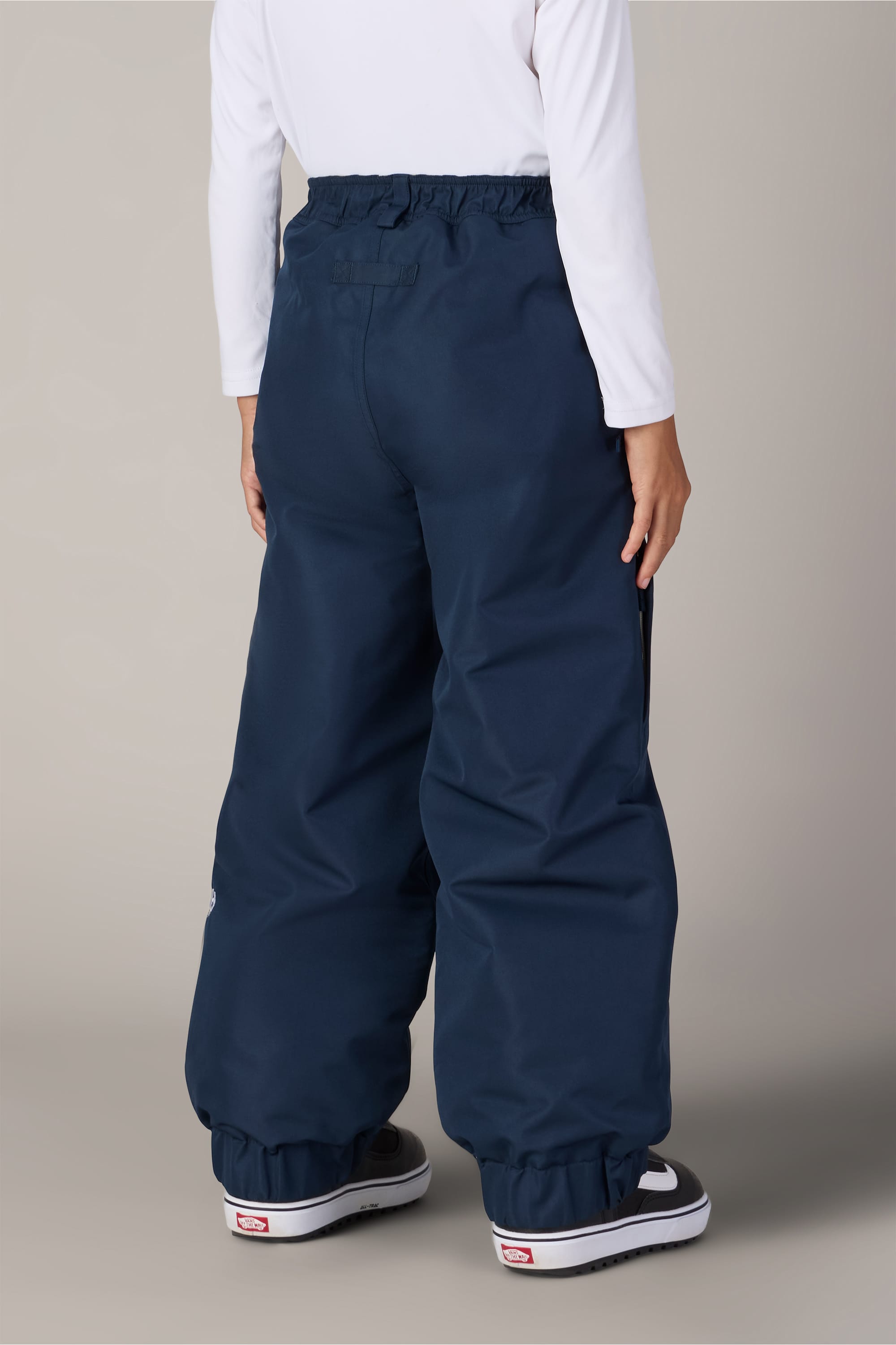 A person wearing the 686 Girls’ Outline™ Insulated Pant in navy blue from 686, paired with a white long-sleeve shirt and black-and-white winter boots, is standing facing away from the camera against a plain light gray background.