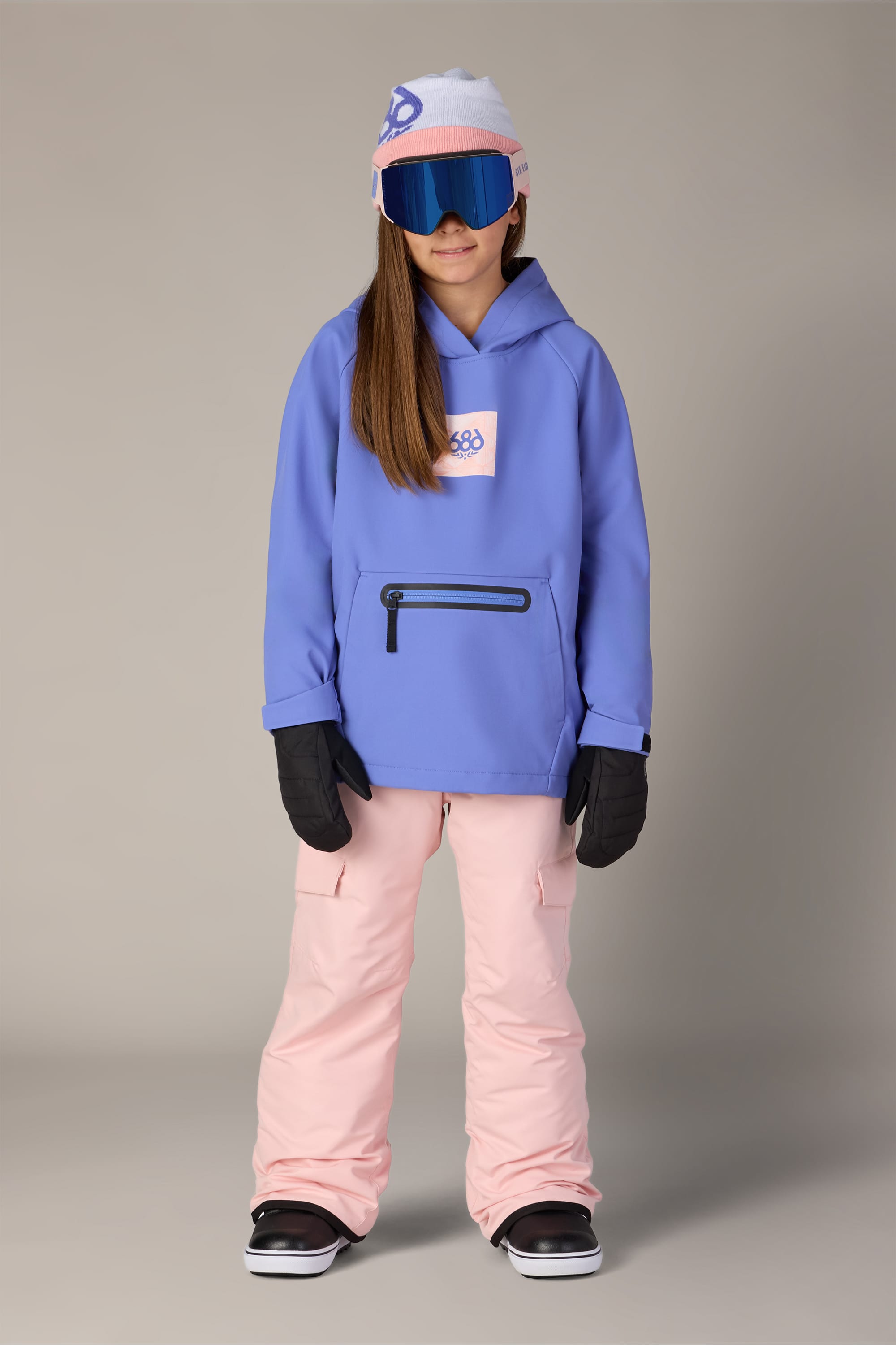 A child wearing a 686 Youth Waterproof Hoody by 686 in light purple, paired with pale pink snow pants, black gloves, blue ski goggles, and a white hat stands facing forward against a plain background.