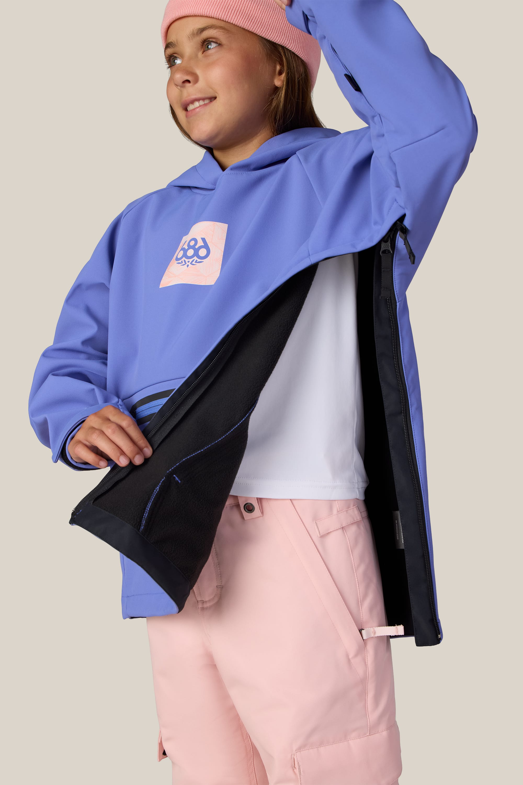 A young girl in a pink beanie, lavender jacket, and pink shorts smiles and looks to the side against a plain background as she opens her 686 Youth Waterproof Hoody to reveal its inner lining.