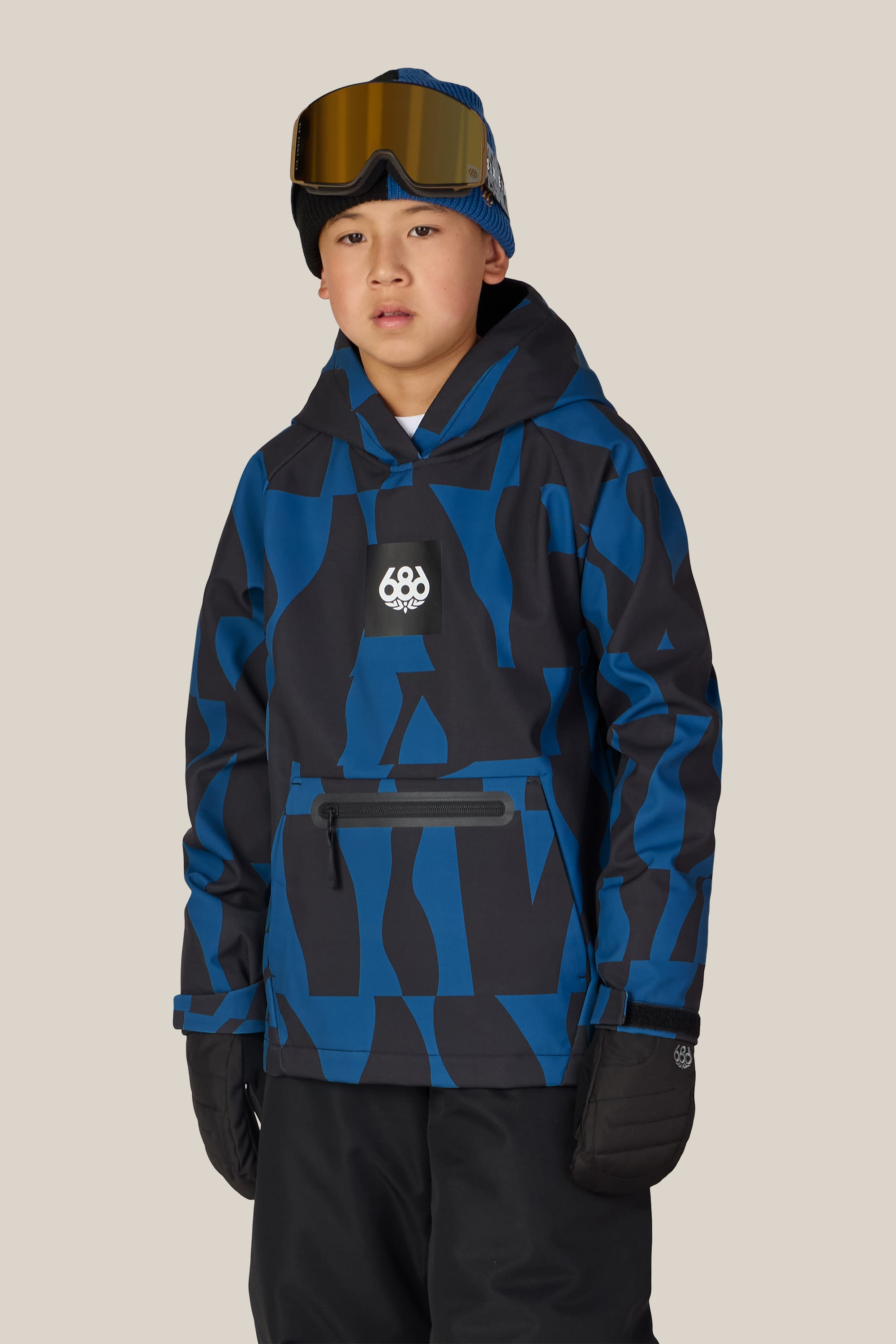 A boy is wearing the 686 Youth Waterproof Hoody by 686, featuring a blue and black pattern, along with black snow pants, black gloves, a blue beanie, and brown ski goggles resting on his forehead. He stands against a plain light background.