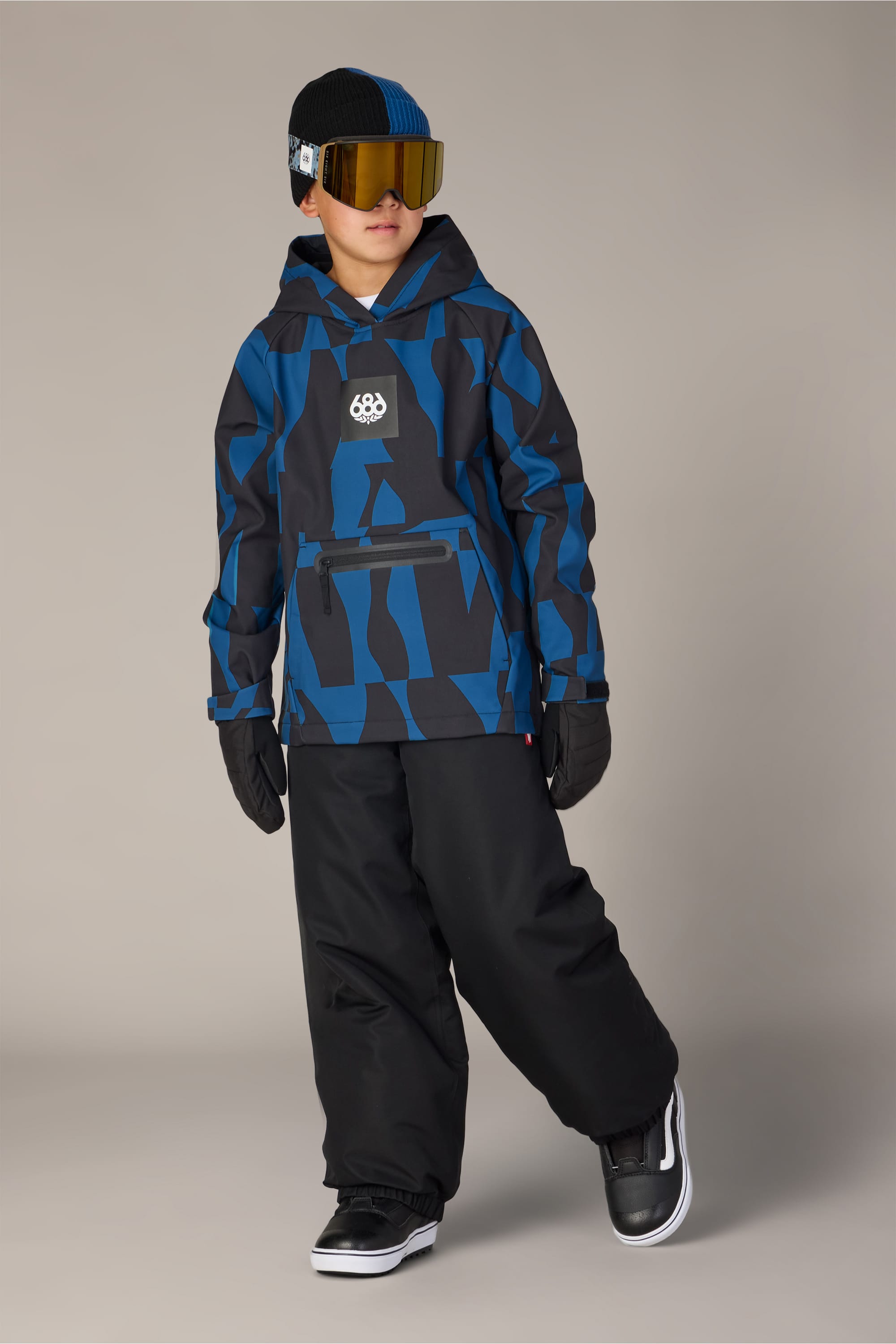 A child wearing a blue and black patterned winter jacket layered over a 686 Youth Waterproof Hoody, along with black snow pants, gloves, black and white sneakers, and large reflective ski goggles, stands against a plain background.