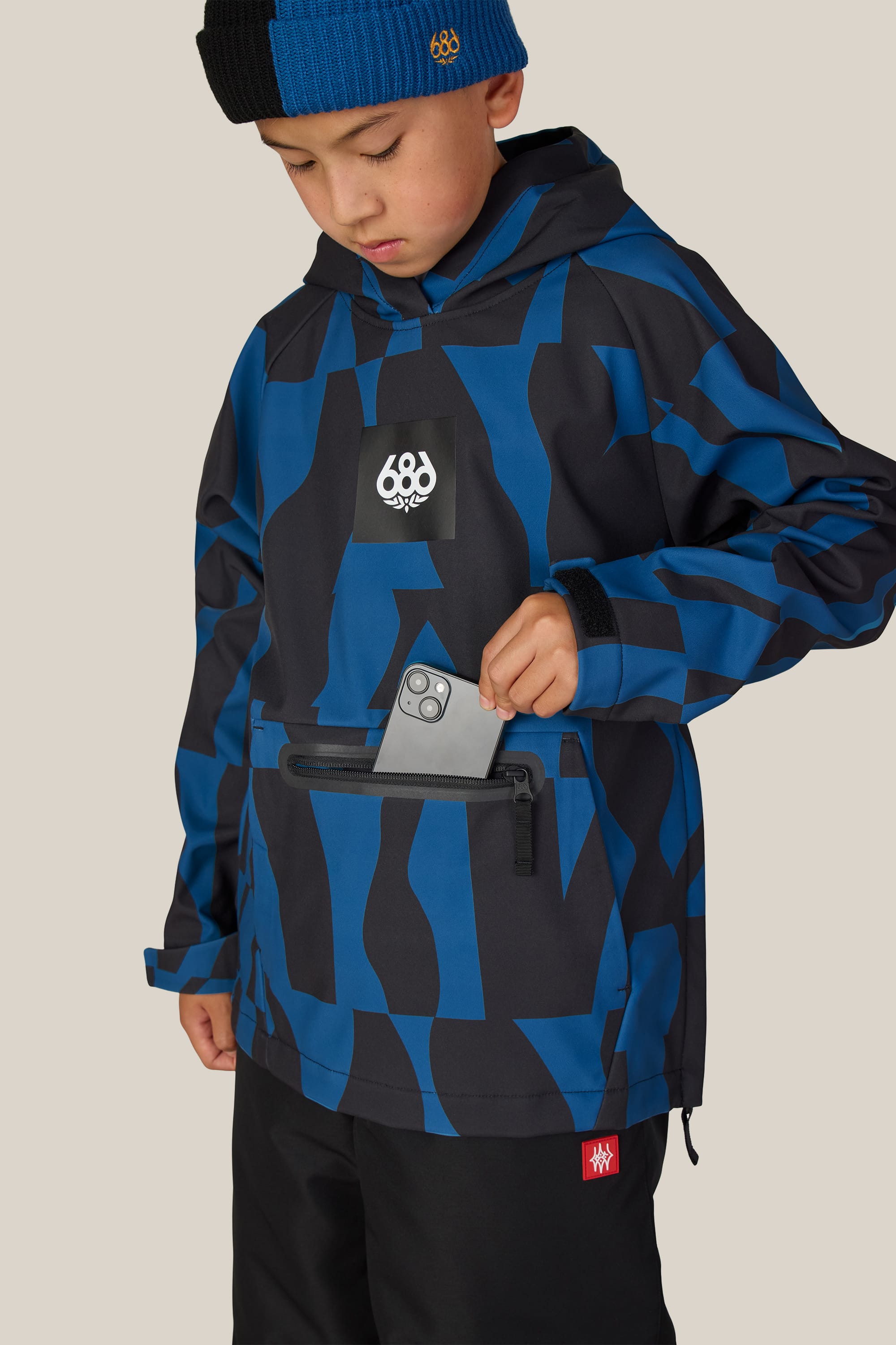 A child wearing the 686 Youth Waterproof Hoody by 686 and a beanie puts a smartphone into the hoody’s front zippered pocket.