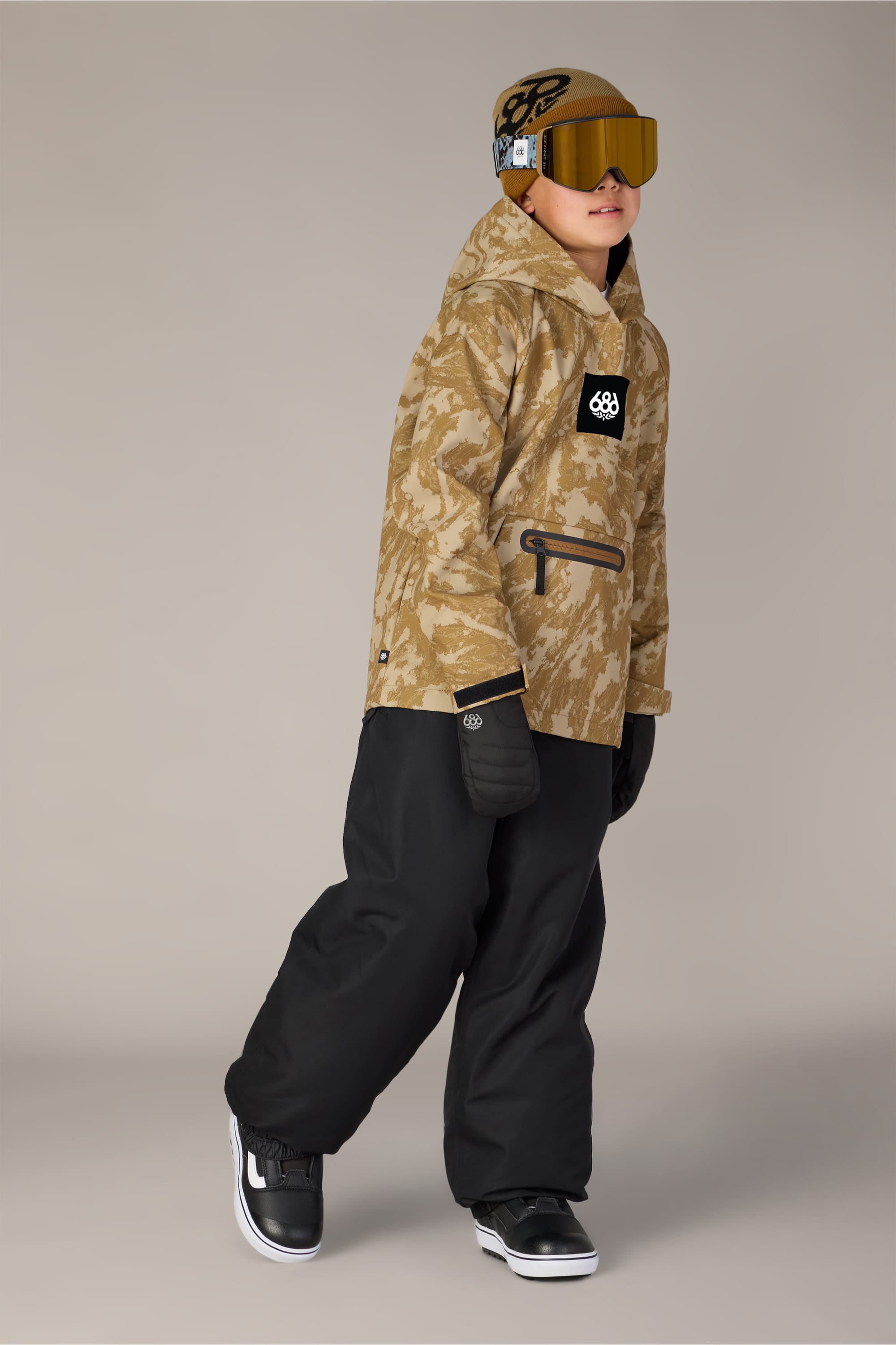 A child wearing a tan camouflage jacket layered over the 686 Youth Waterproof Hoody by 686, black snow pants, black gloves, black and white sneakers, and a tan helmet with goggles stands against a plain background.
