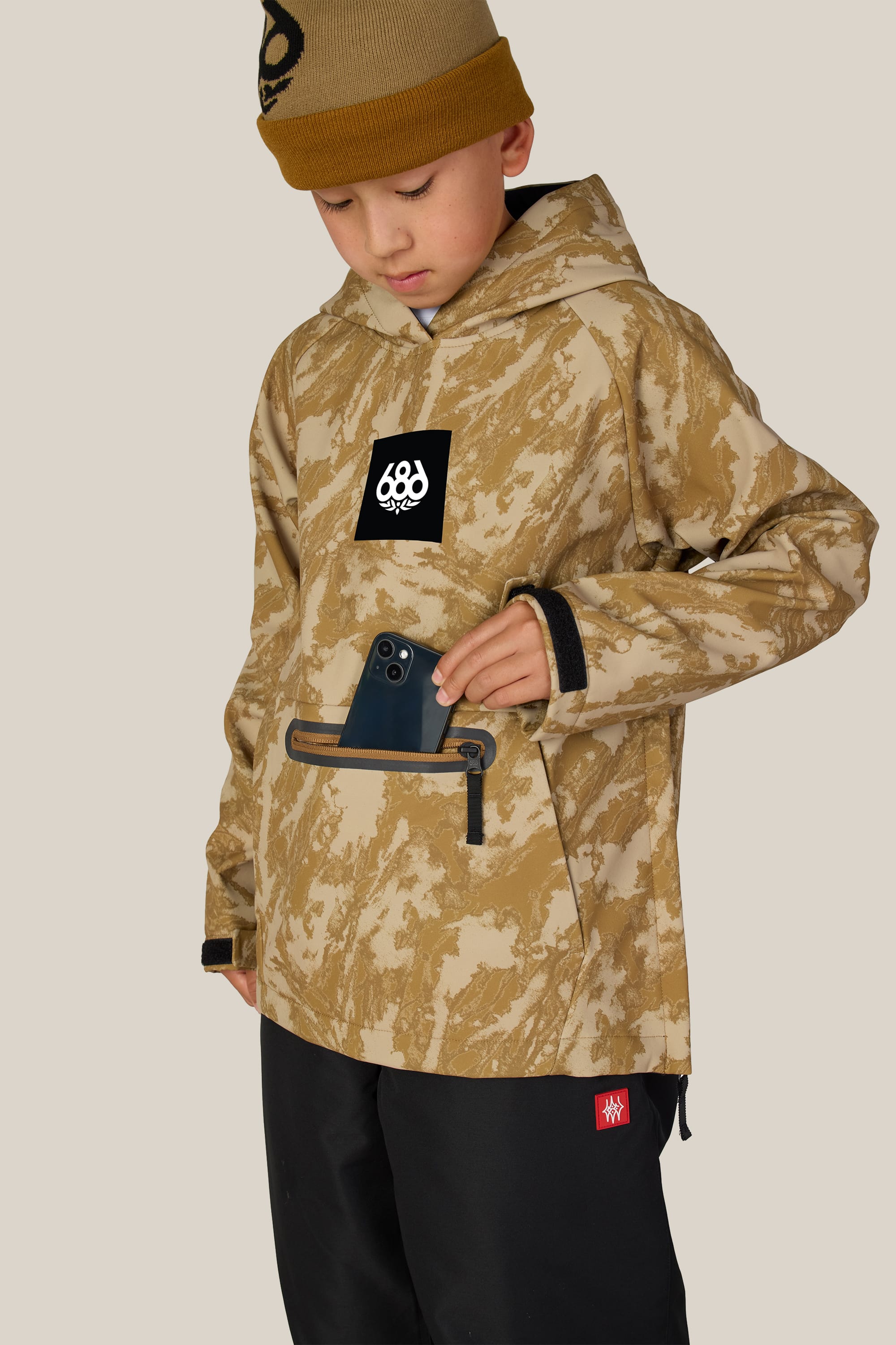 A young boy wearing the 686 Youth Waterproof Hoody in tan camo and a brown beanie places a black smartphone into the front zip pocket while looking down. The hoody, by 686, features a black logo on the chest. The background is plain beige.