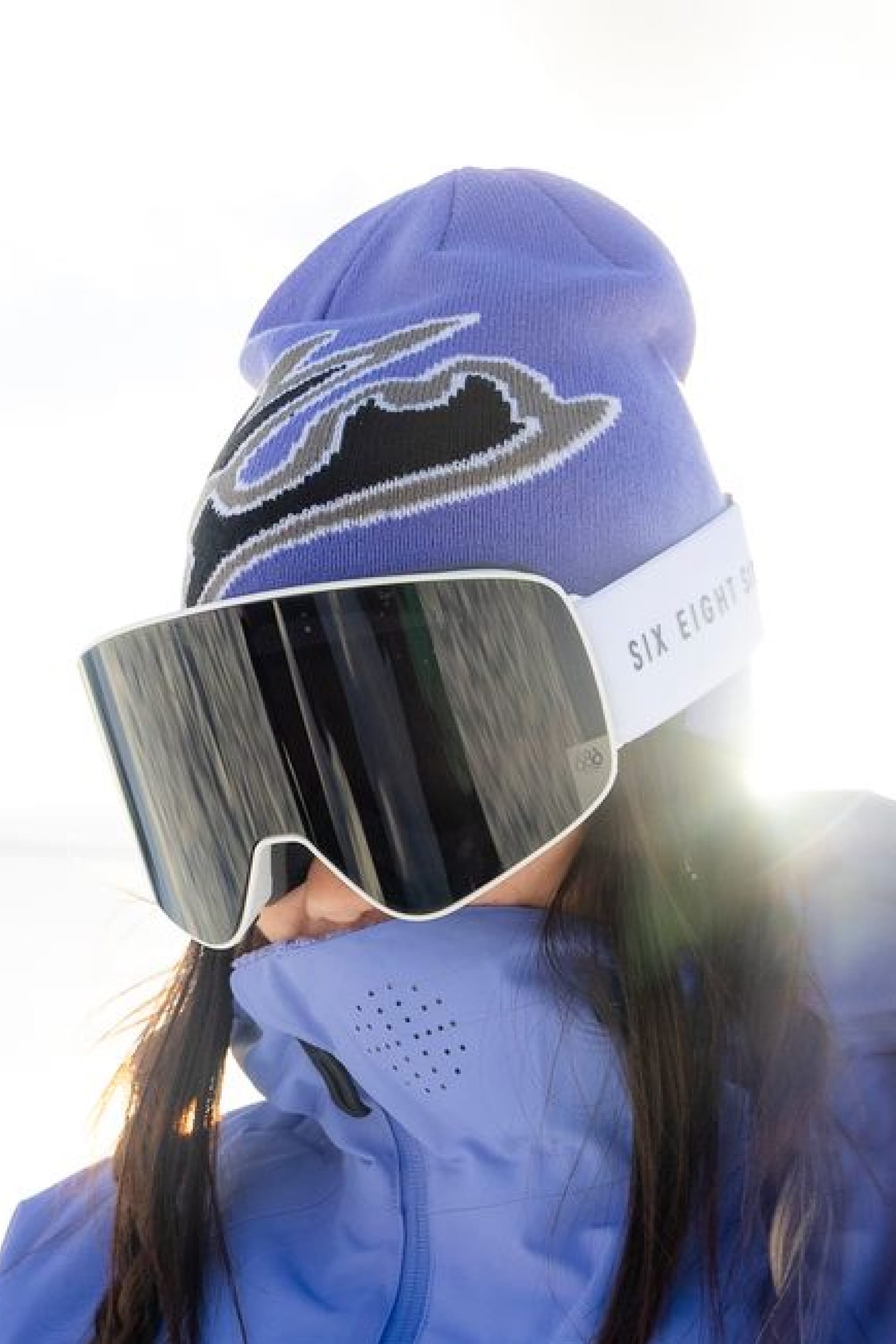 A person in a purple jacket and the 686 Jib Skully Beanie—a matching acrylic knit beanie by 686 with a black and white design—along with large reflective ski goggles stands outdoors, facing sunlight.