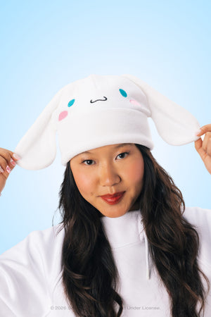 A woman wearing the 686 Women’s Cinnamoroll™ Beanie holds its ear flaps and smiles against a light blue background.