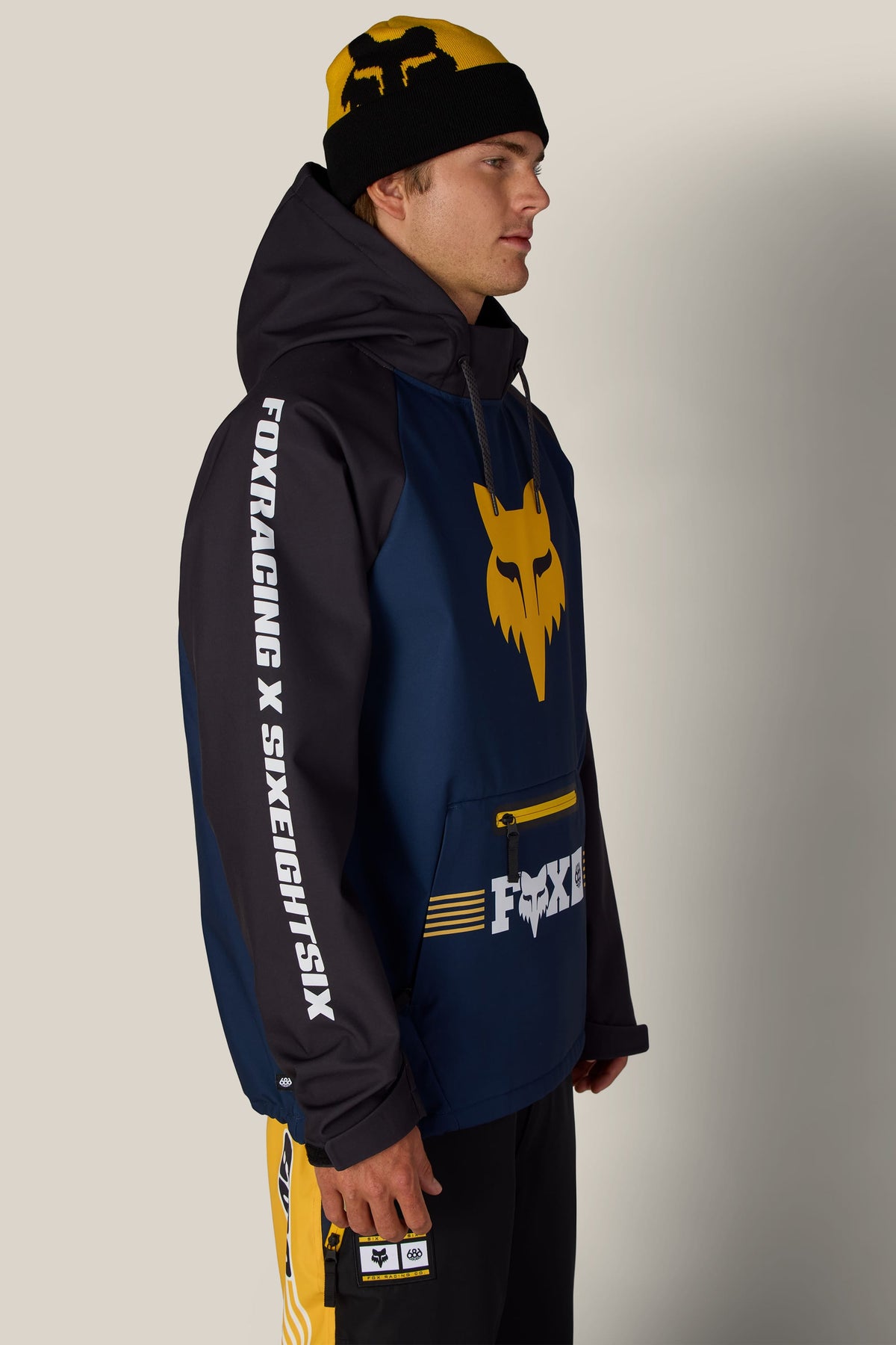A man in profile wears a 686 Men’s Fox Racing Waterproof Hoody, a matching yellow and black beanie, and black pants.