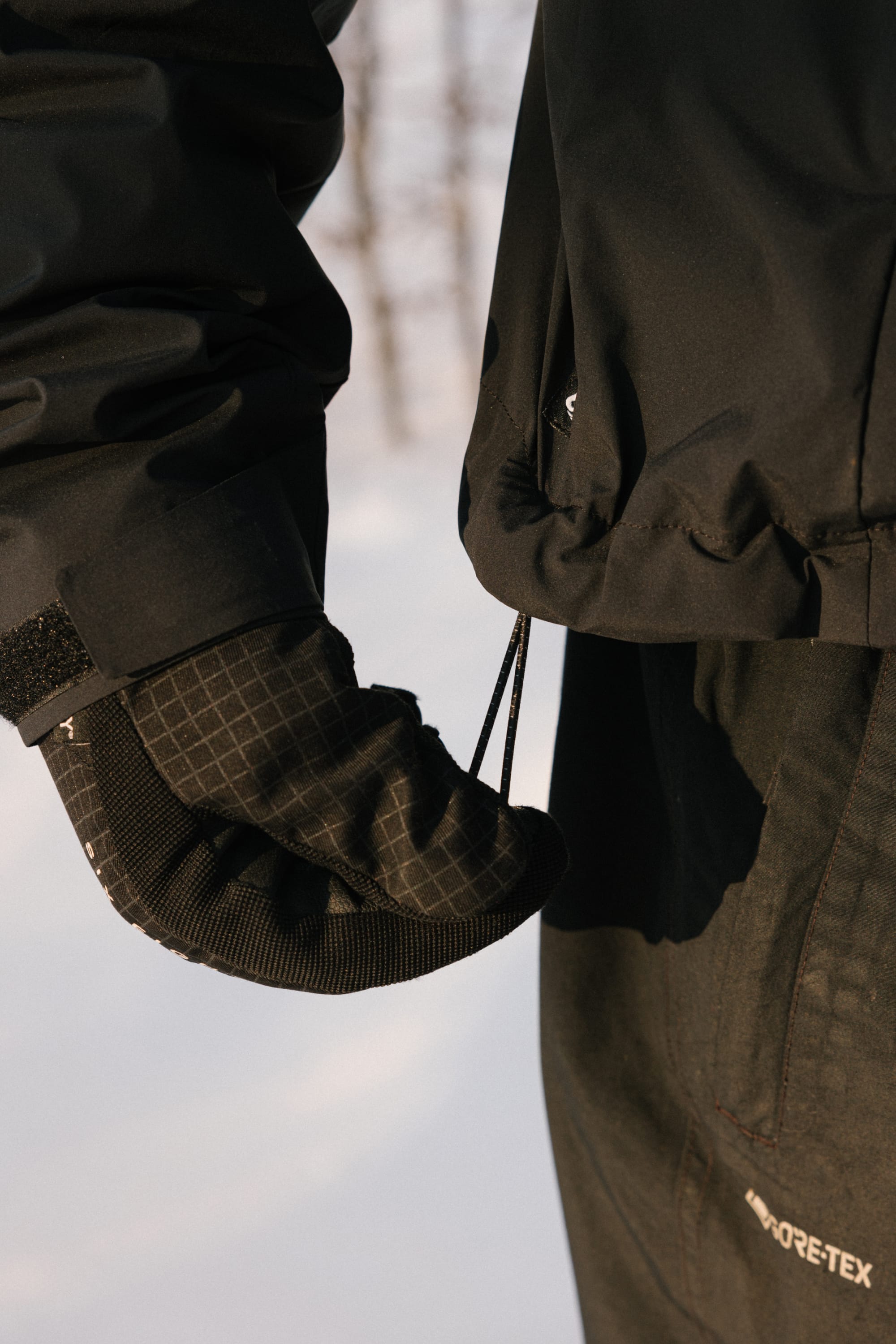 A person wearing a black winter glove and jacket, possibly the 686 Men’s WINDSTOPPER® Pullover Hoody by GORE-TEX® LABS from 686, is pulling a drawstring on their coat while snow falls in the blurred background.