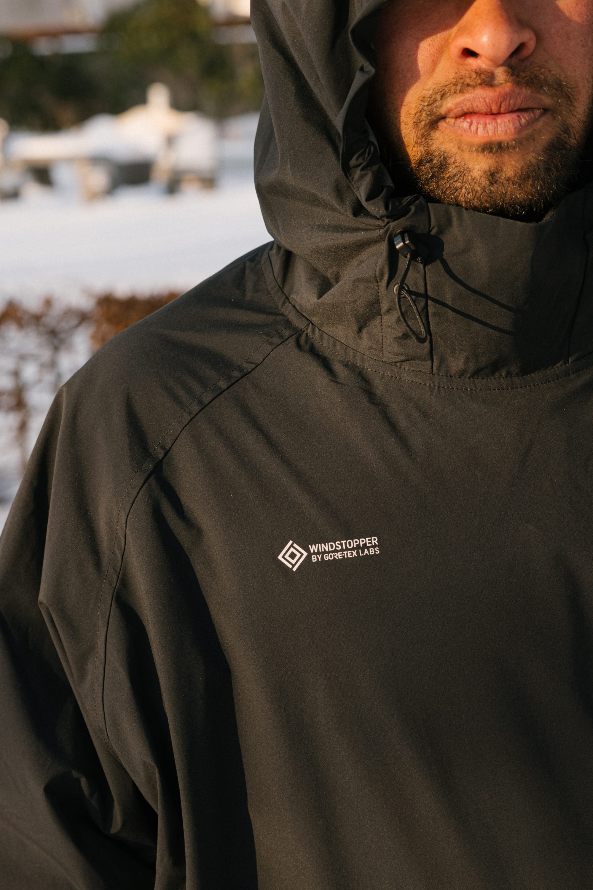 A person stands outdoors in a snowy area wearing a black 686 Men’s WINDSTOPPER® Pullover Hoody by GORE-TEX® LABS, with the hood up. The focus is on the jacket’s logo, and the person’s face is partially visible.