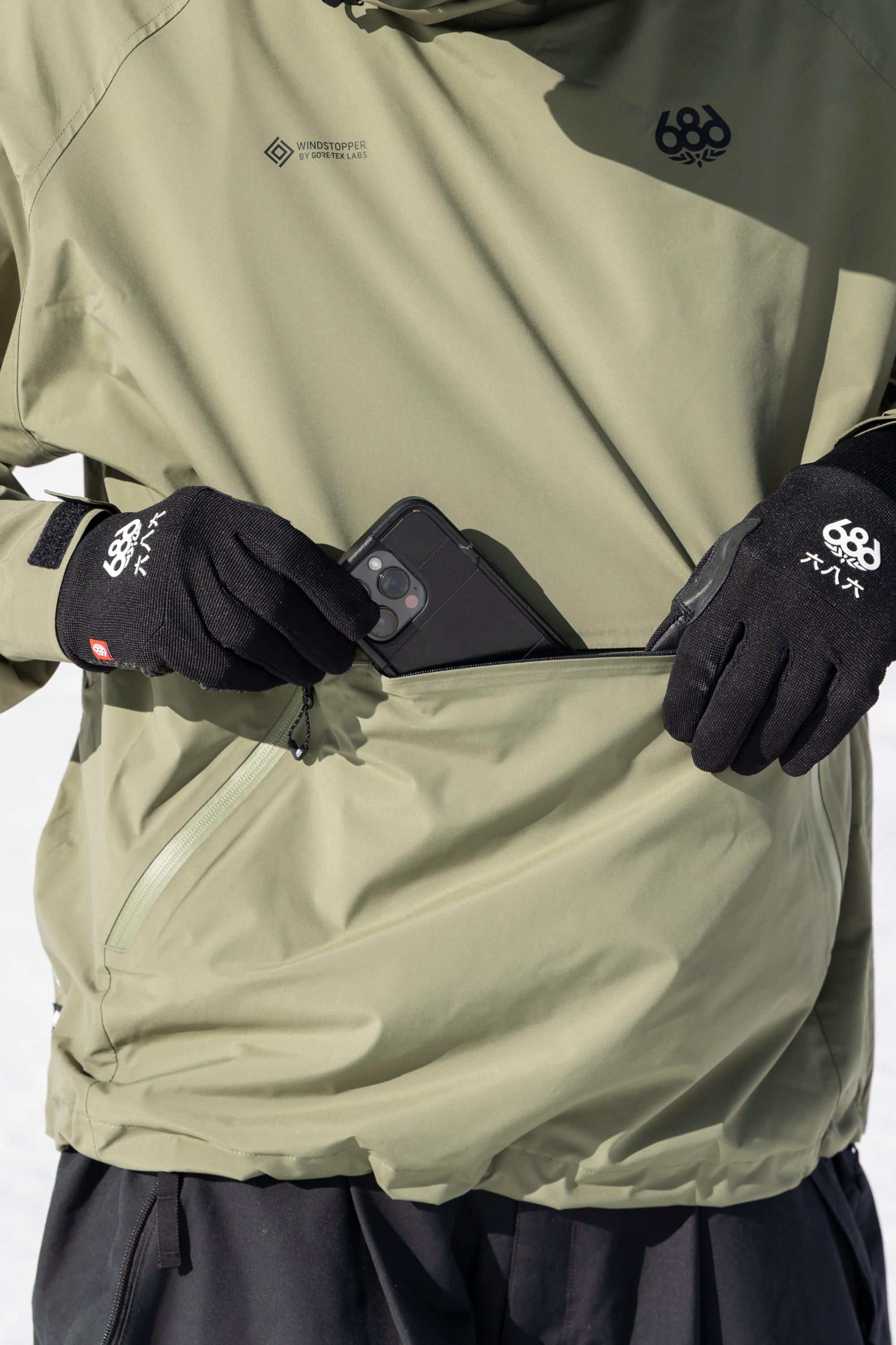 Someone, clad in black gloves and the 686 Men’s WINDSTOPPER® Pullover Hoody by GORE-TEX® LABS, zips a black smartphone into the front pocket. Only their torso and hands are visible against a snowy backdrop.