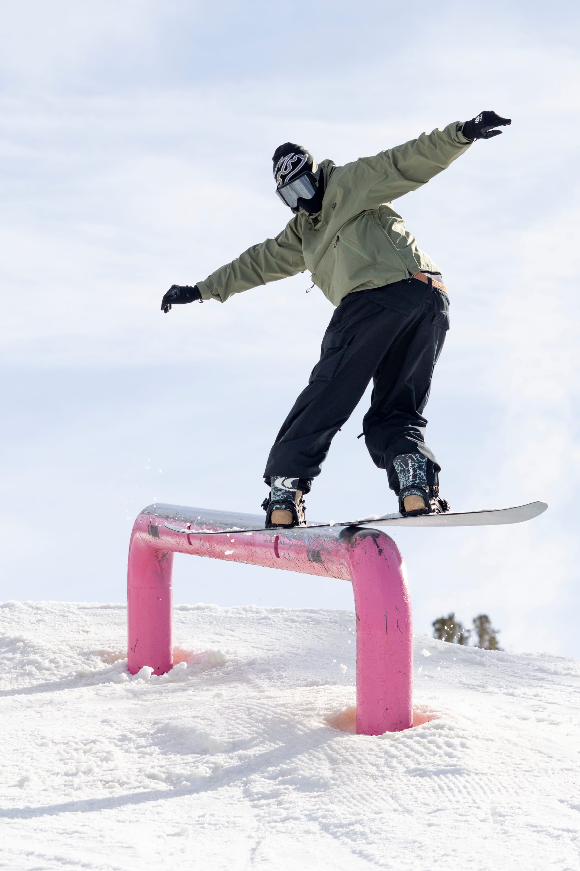 A snowboarder, dressed in a 686 Men’s WINDSTOPPER® Pullover Hoody by GORE-TEX® LABS beneath his green jacket, balances on a pink rail in the snow. He pairs black pants, gloves, and a helmet with goggles against the backdrop of a bright, cloudy sky.