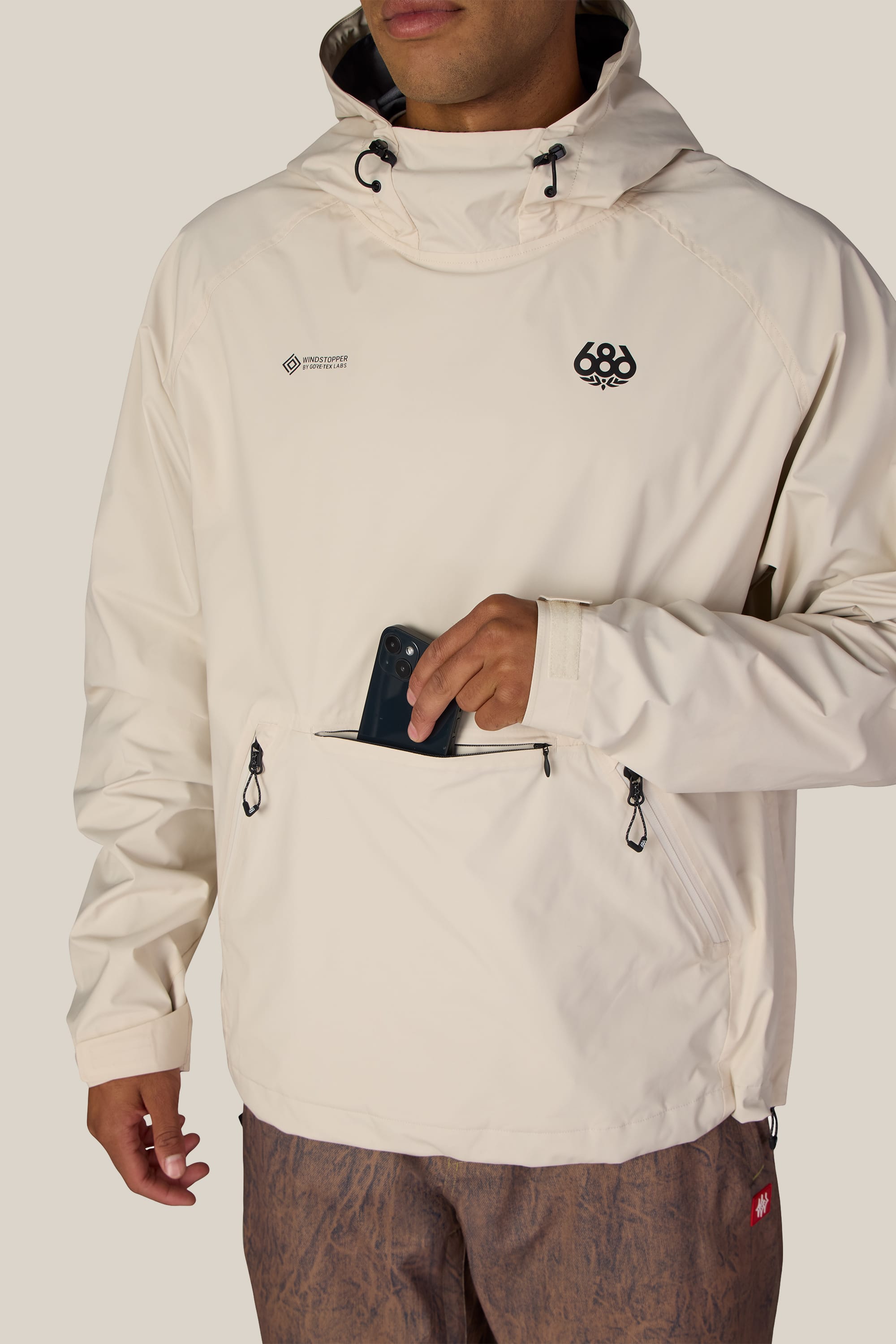 A person wearing the 686 Men’s WINDSTOPPER® Pullover Hoody by GORE-TEX® LABS—a cream polyester hoody with front zippers—places a phone into the center pocket. The woven jacket features two small black 686 logos on the chest, and only the lower half of the face is visible.