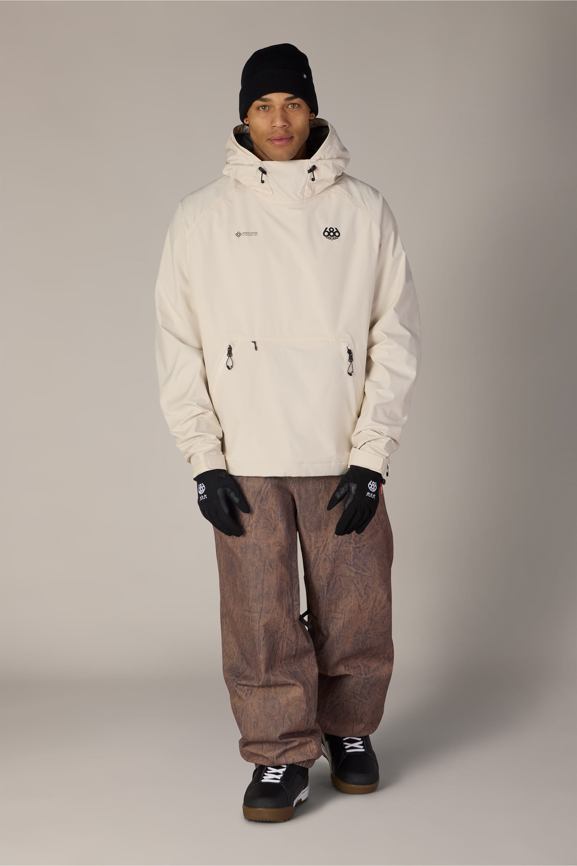 A person wearing the 686 Men’s WINDSTOPPER® Pullover Hoody by GORE-TEX® LABS in cream, styled with a black beanie, black gloves, loose brown pants, and black shoes, stands against a plain light grey background.