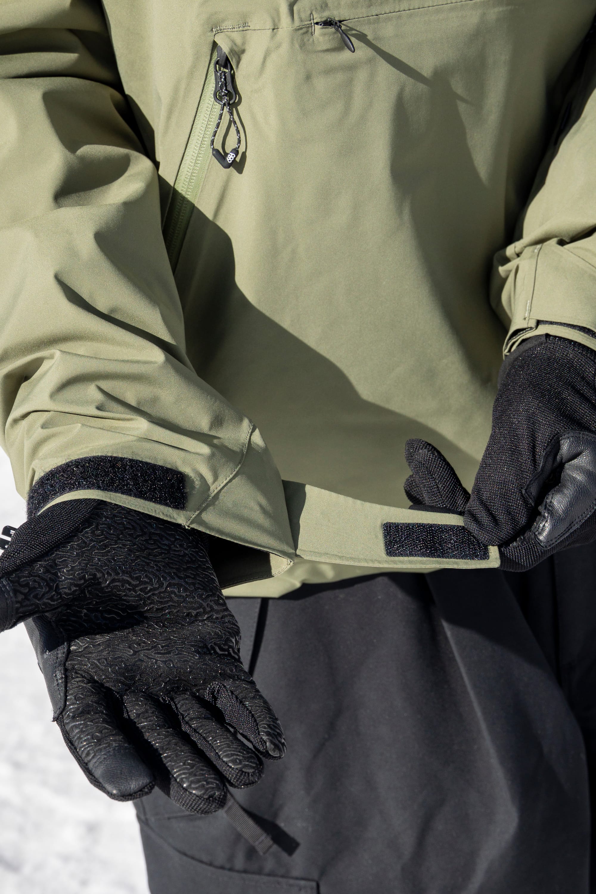 Wearing black gloves and the 686 Men’s WINDSTOPPER® Pullover Hoody by GORE-TEX® LABS, a person adjusts the Velcro strap on their sleeve while standing outdoors in a snowy environment.