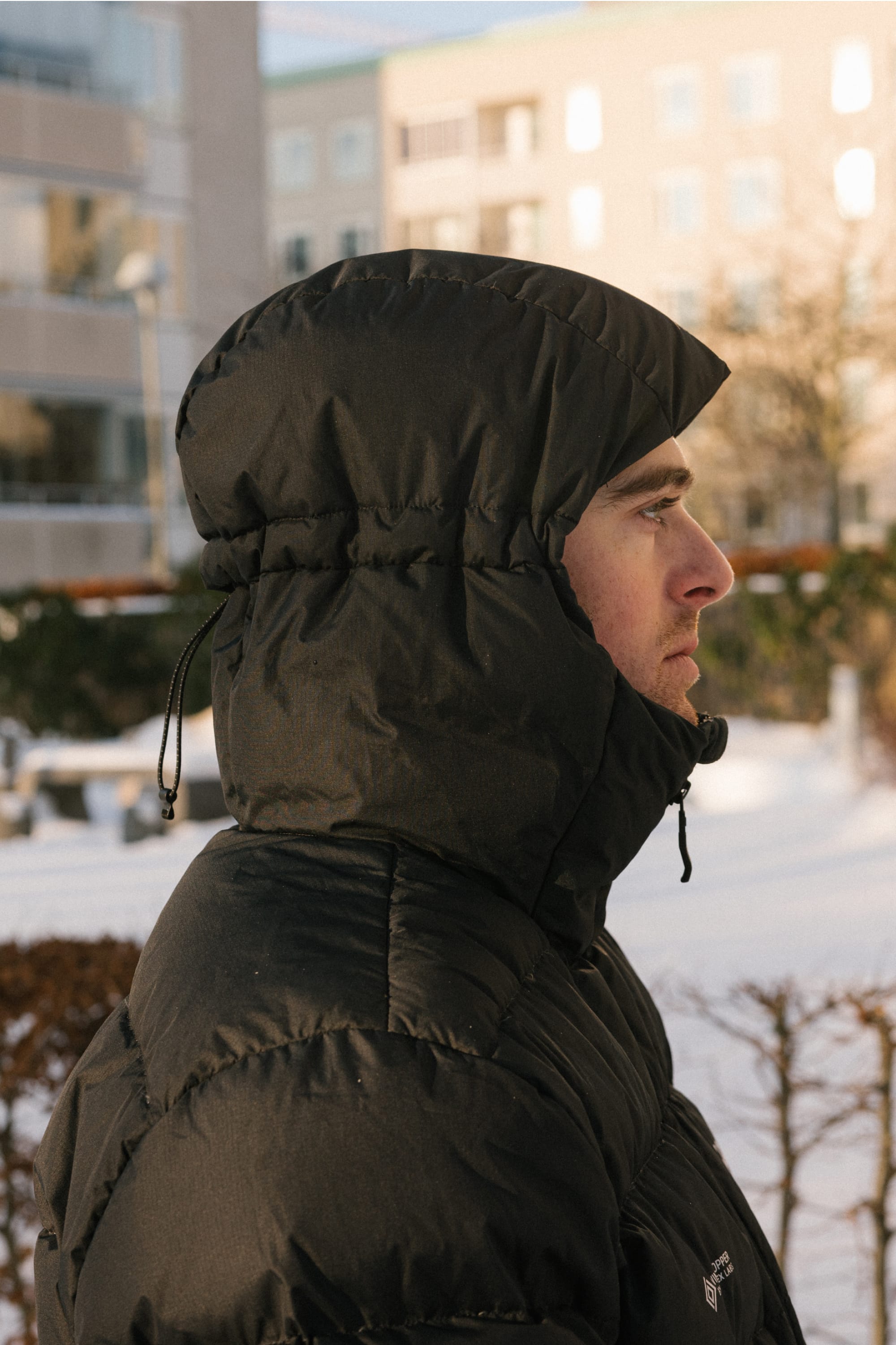 A person in profile stands outside, with snow on the ground and buildings in the background, wearing a black 686 Men’s WINDSTOPPER® Pyro™ Down Puffer Jacket by GORE-TEX® LABS.