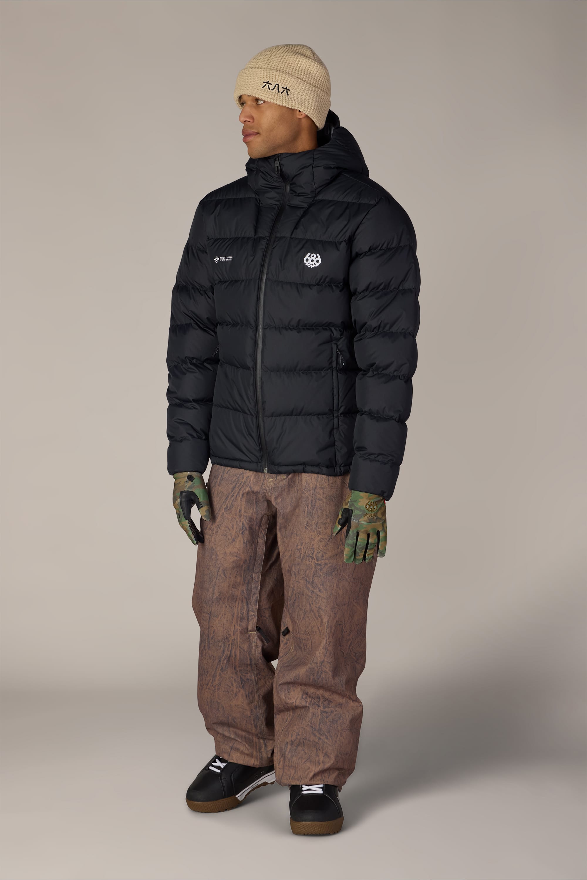 A person wearing a beige beanie, the 686 Men’s WINDSTOPPER® Pyro™ Down Puffer Jacket by GORE-TEX® LABS in black, brown baggy pants, green gloves, and black sneakers stands facing slightly right against a neutral background.