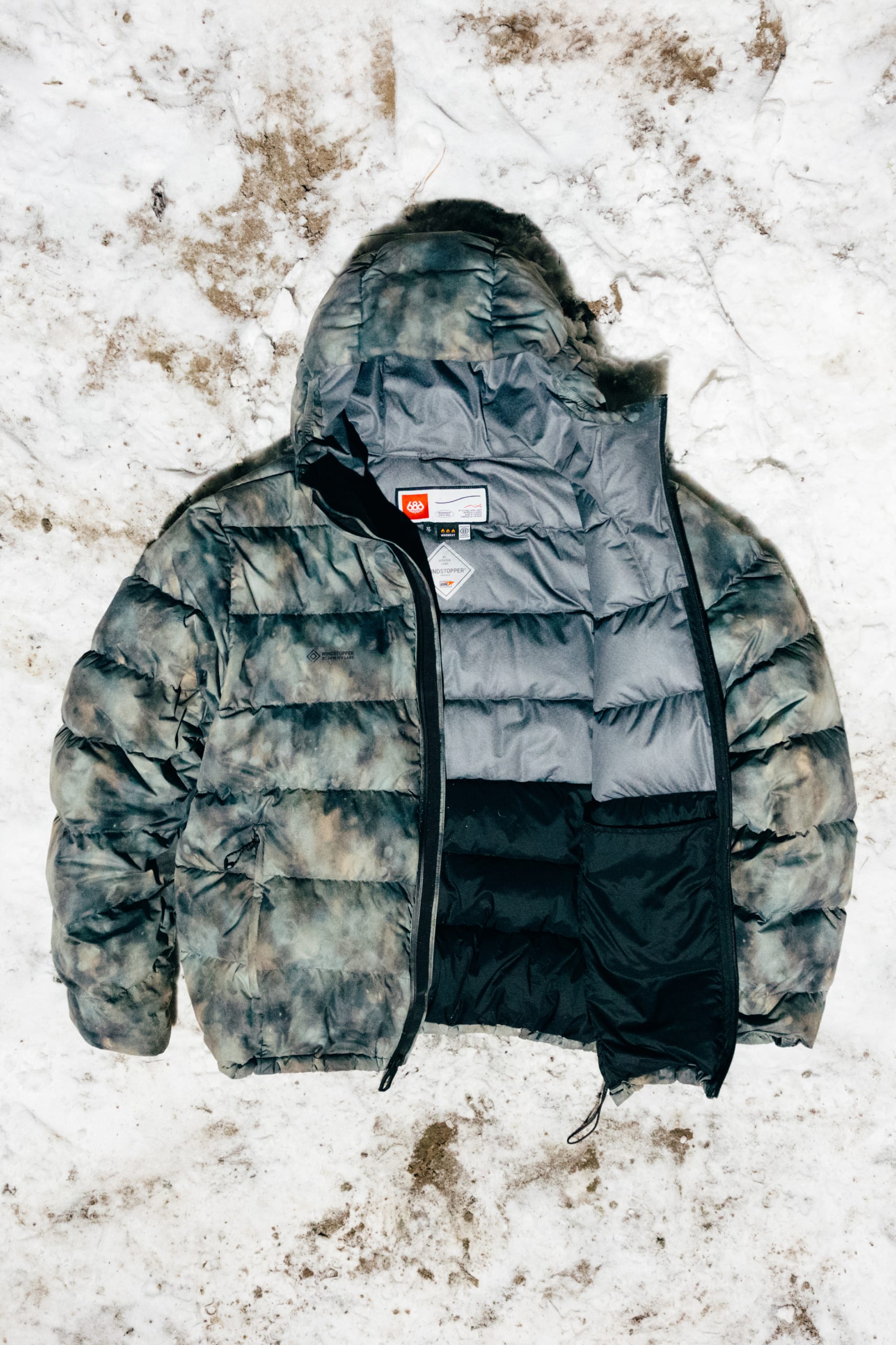 The 686 Men’s WINDSTOPPER® Pyro™ Down Puffer Jacket by GORE-TEX® LABS, featuring a camouflage pattern and hooded puffer design, is laid open on snowy ground to display its gray and black quilted polyester lining and visible collar tags.