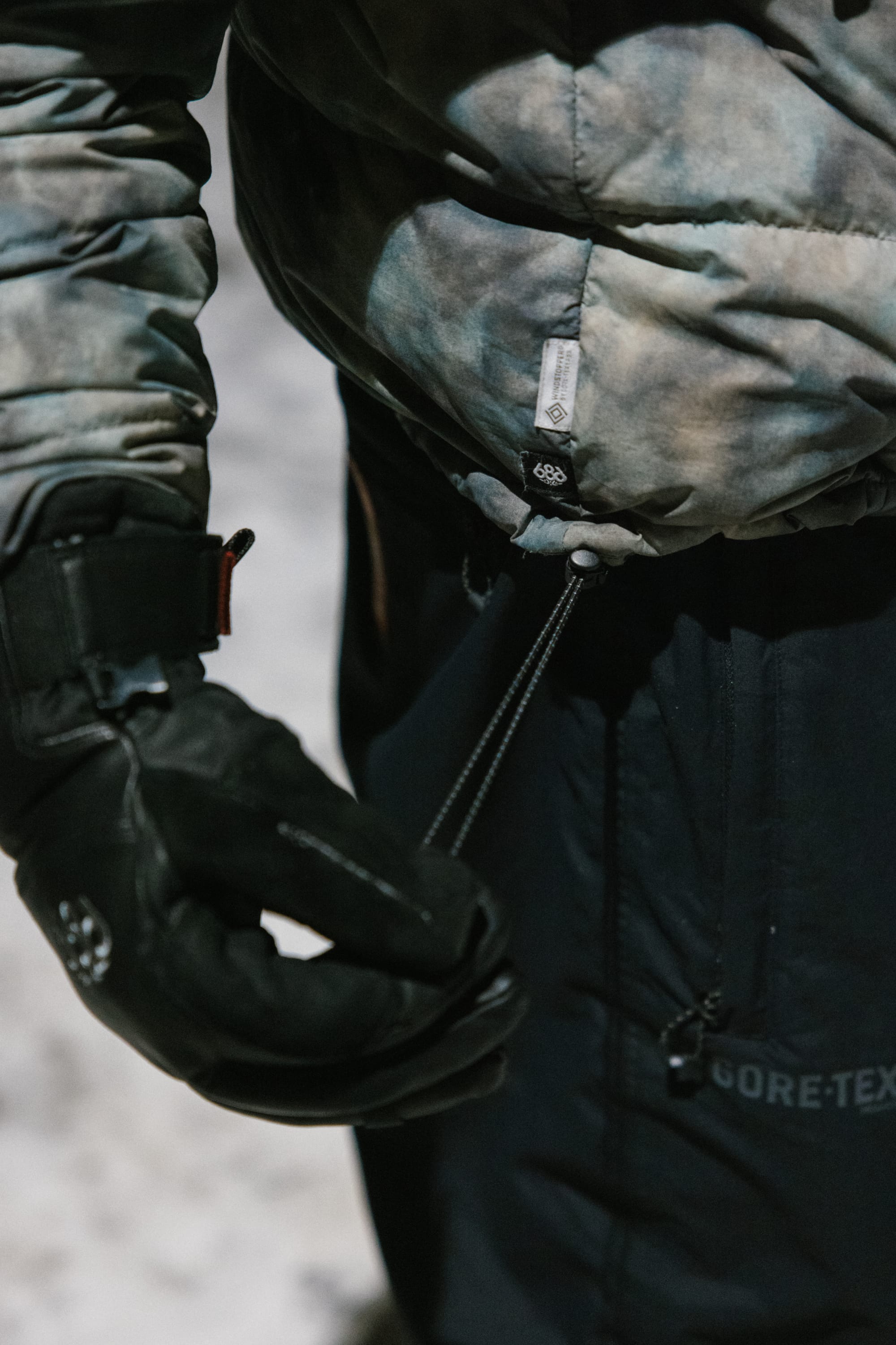 A person wearing black gloves and a 686 Men’s WINDSTOPPER® Pyro™ Down Puffer Jacket by GORE-TEX® LABS tightens the jacket’s waist drawstring. The word GORE-TEX is visible on the pants, with blurred snow in the background.