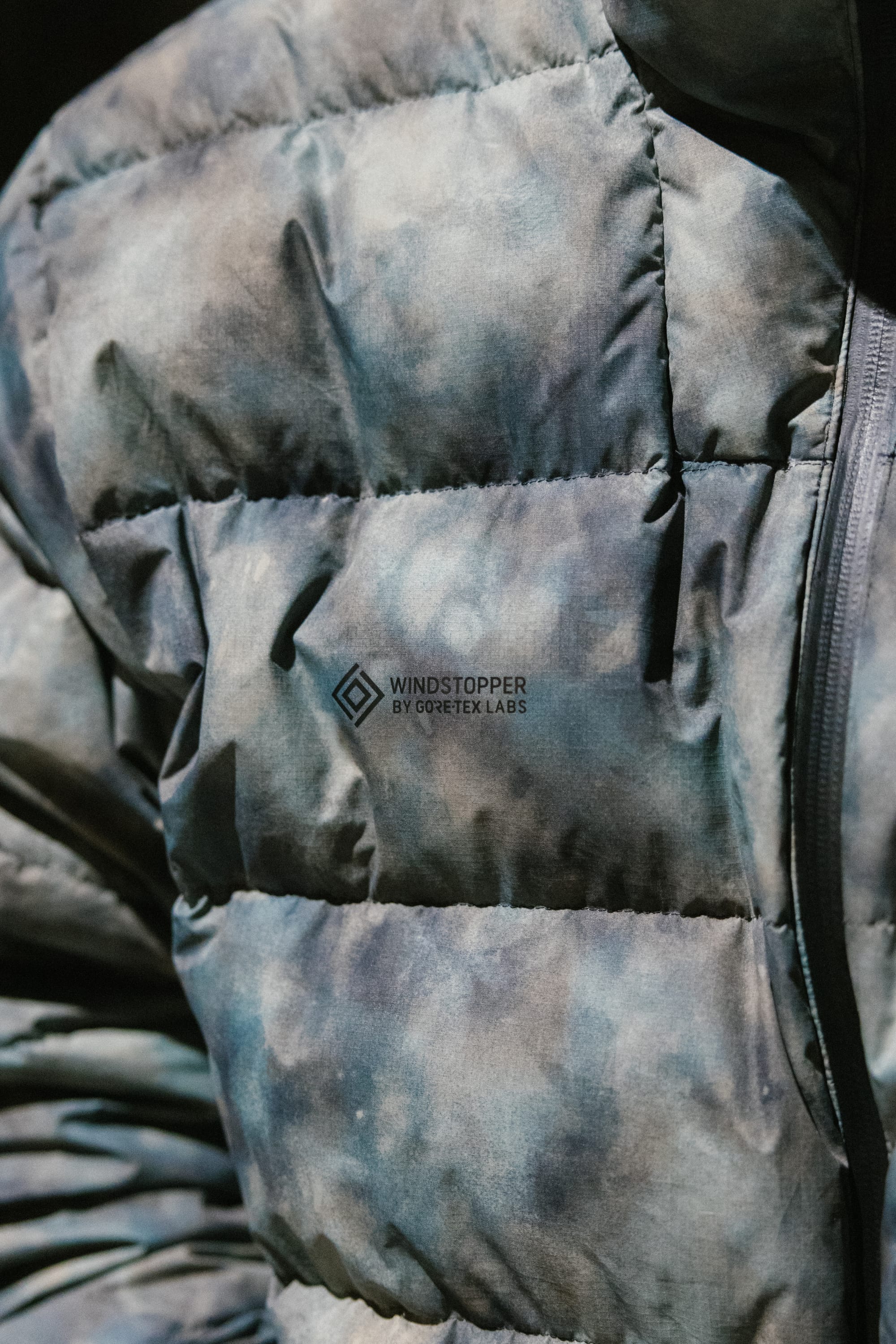 A close-up of the 686 Men’s WINDSTOPPER® Pyro™ Down Puffer Jacket by GORE-TEX® LABS, featuring a blue and gray marbled design with the WINDSTOPPER BY GORE-TEX LABS logo printed on the chest.