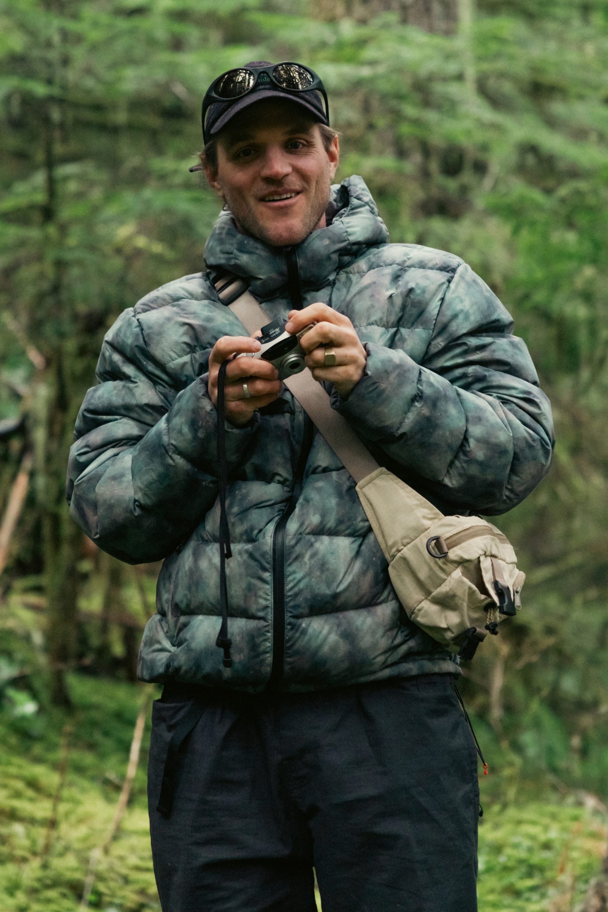 A person in the 686 Men’s WINDSTOPPER® Pyro™ Down Puffer Jacket by GORE-TEX® LABS stands outdoors in a forest, holding a camera and smiling, with a cap, beige crossbody bag, and sunglasses on their head.