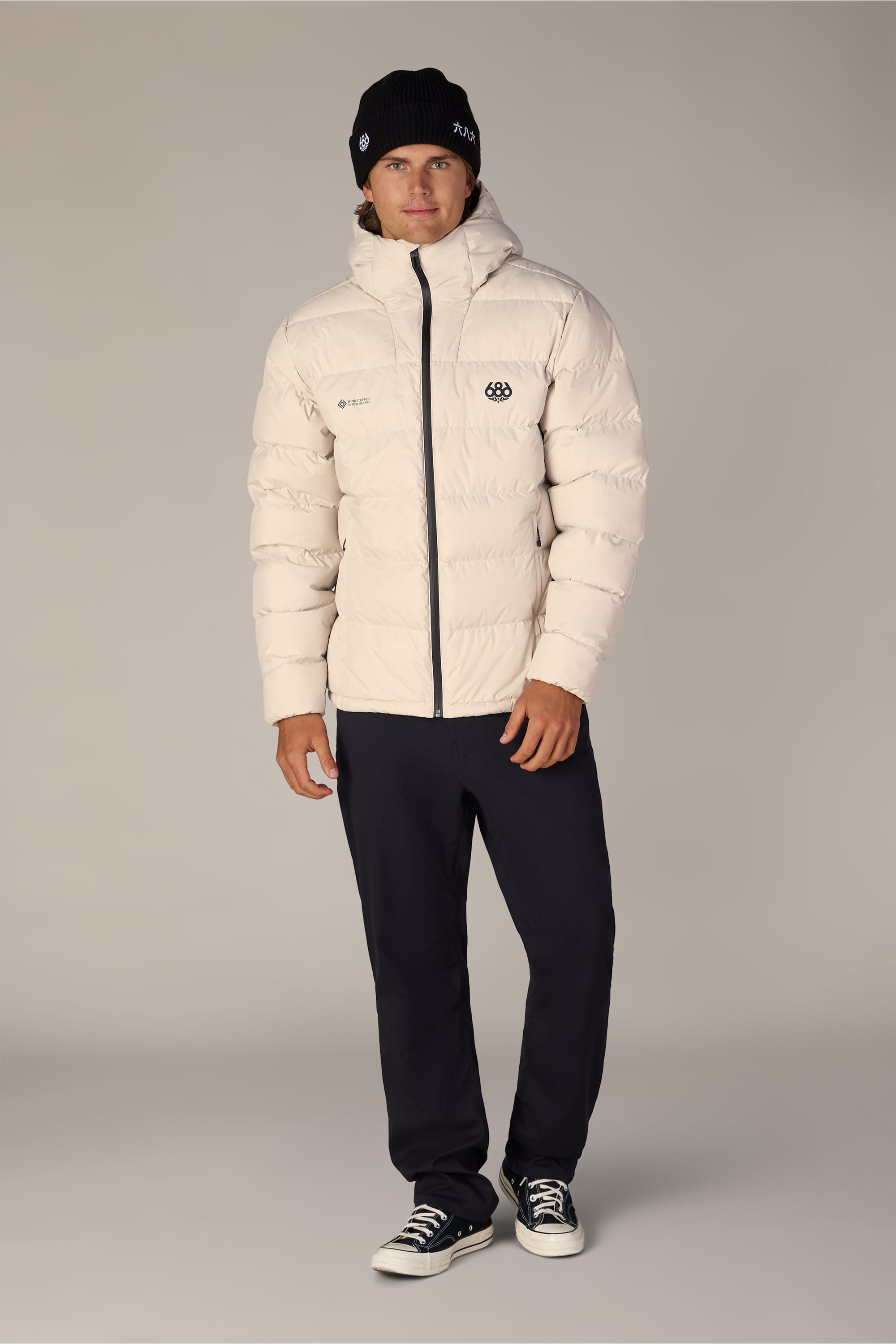 A person wearing the 686 Men’s WINDSTOPPER® Pyro™ Down Puffer Jacket by GORE-TEX® LABS, styled in white and paired with a black beanie, black pants, and black-and-white sneakers, stands against a plain grey background.