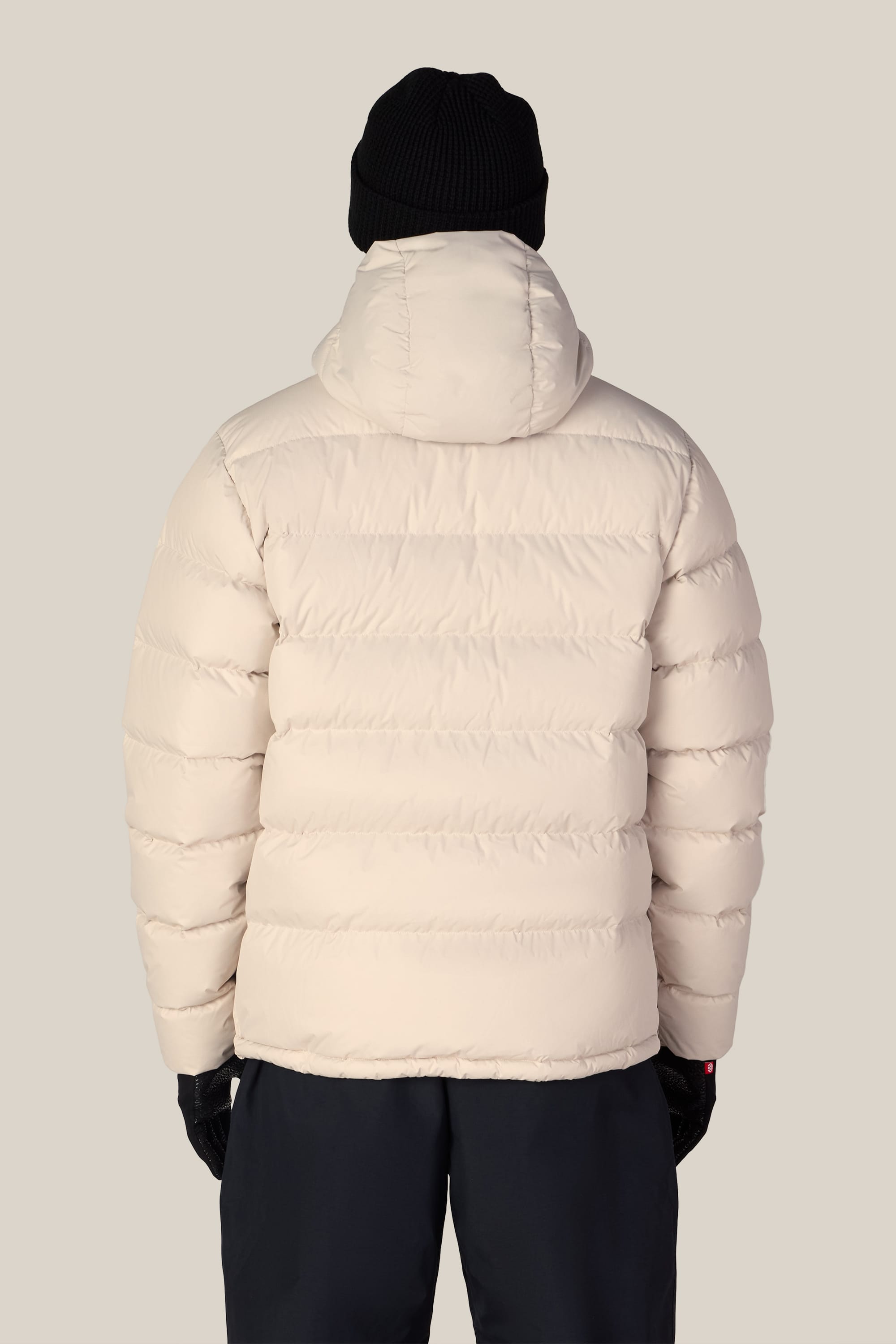 A person is seen from behind against a plain light background, wearing the 686 Men’s WINDSTOPPER® Pyro™ Down Puffer Jacket by GORE-TEX® LABS in a cream color, paired with a black beanie, black gloves, and dark pants.