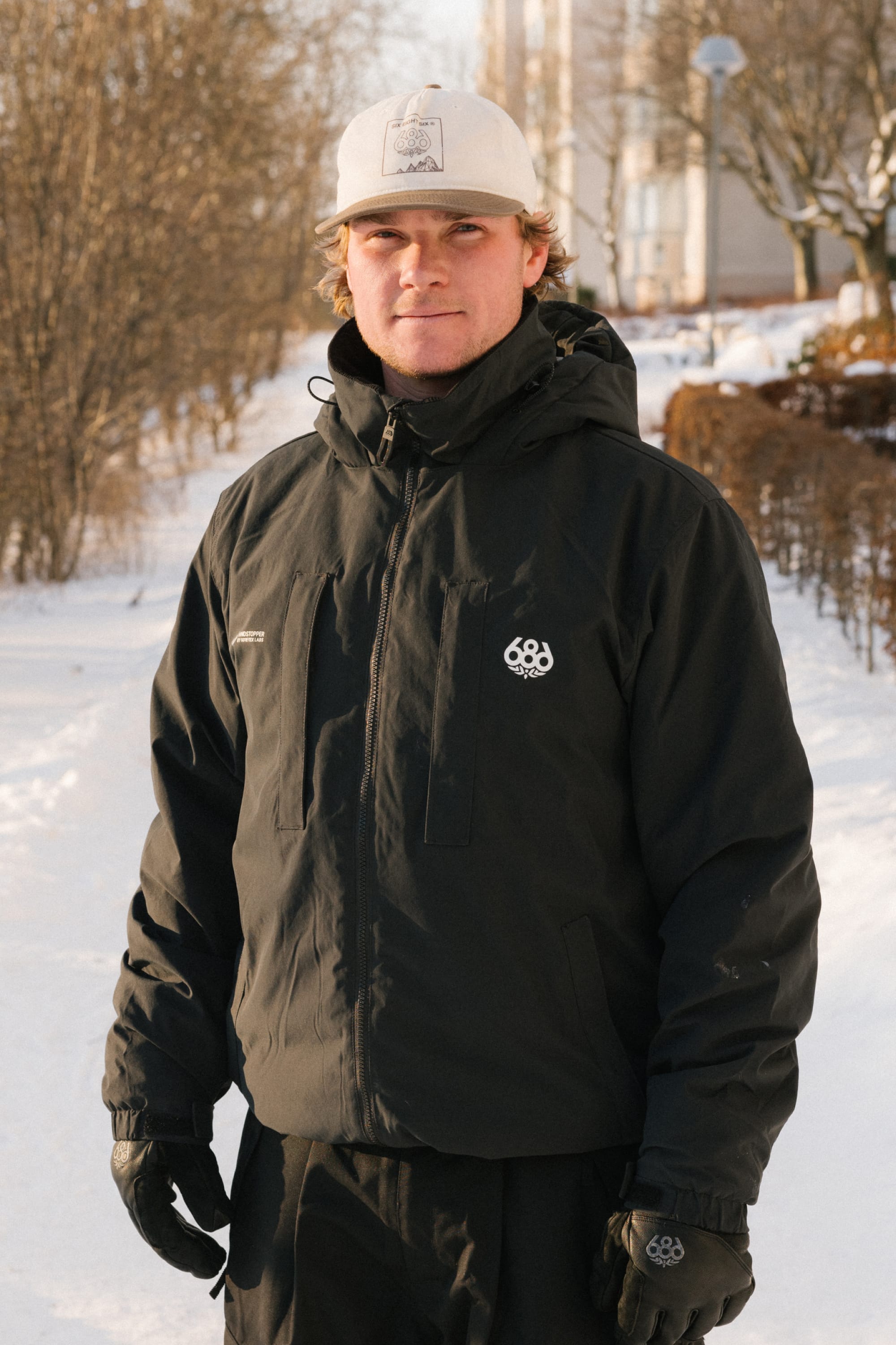 A person wearing the 686 Men’s WINDSTOPPER® Guideline Insulated Jacket by GORE-TEX® LABS stands outdoors on a snowy path, dressed in black gloves and a white cap, with bare trees and buildings visible in the background.