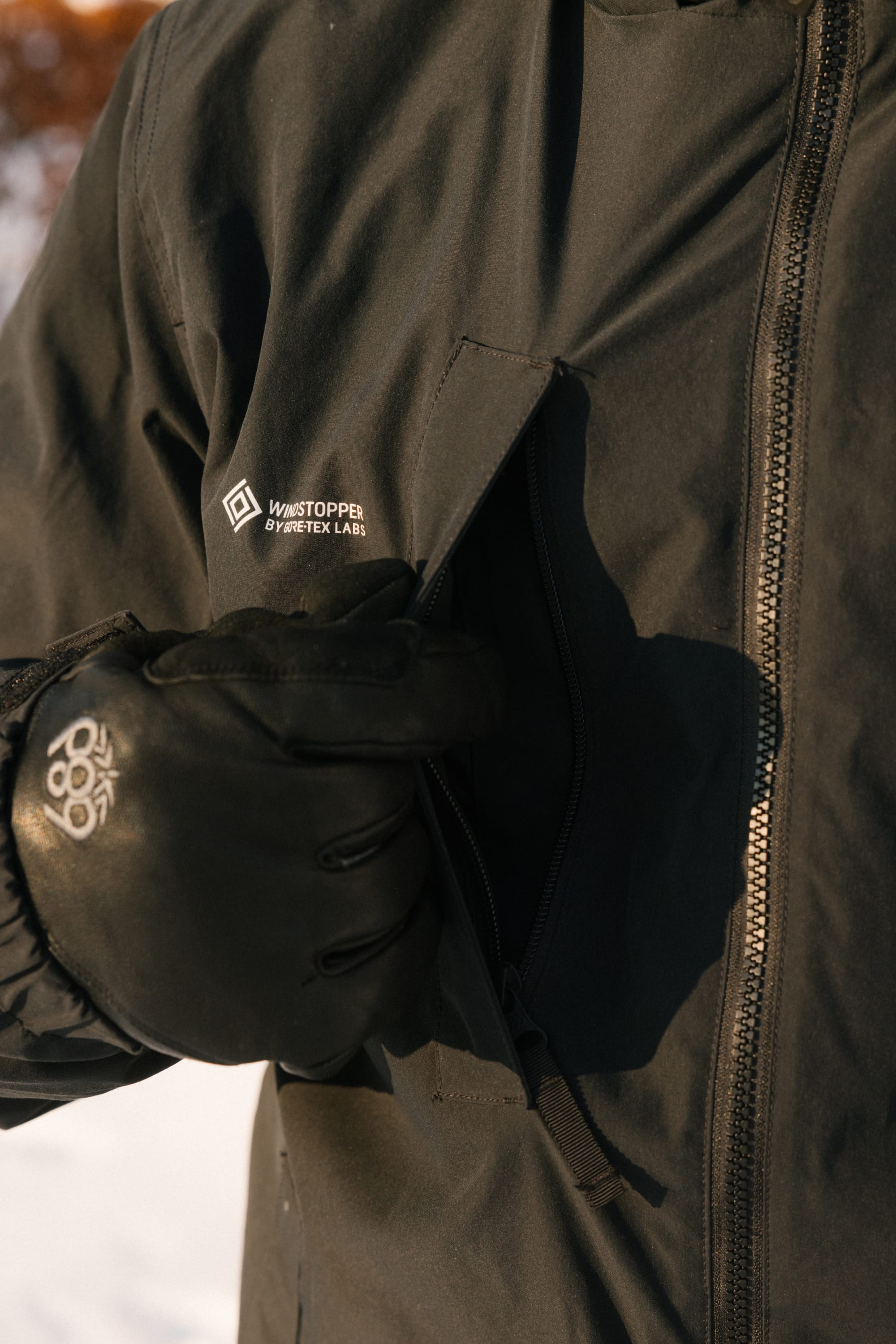 A person wearing black gloves and the 686 Men’s WINDSTOPPER® Guideline Insulated Jacket by GORE-TEX® LABS opens a zippered chest pocket outdoors in a snowy setting.