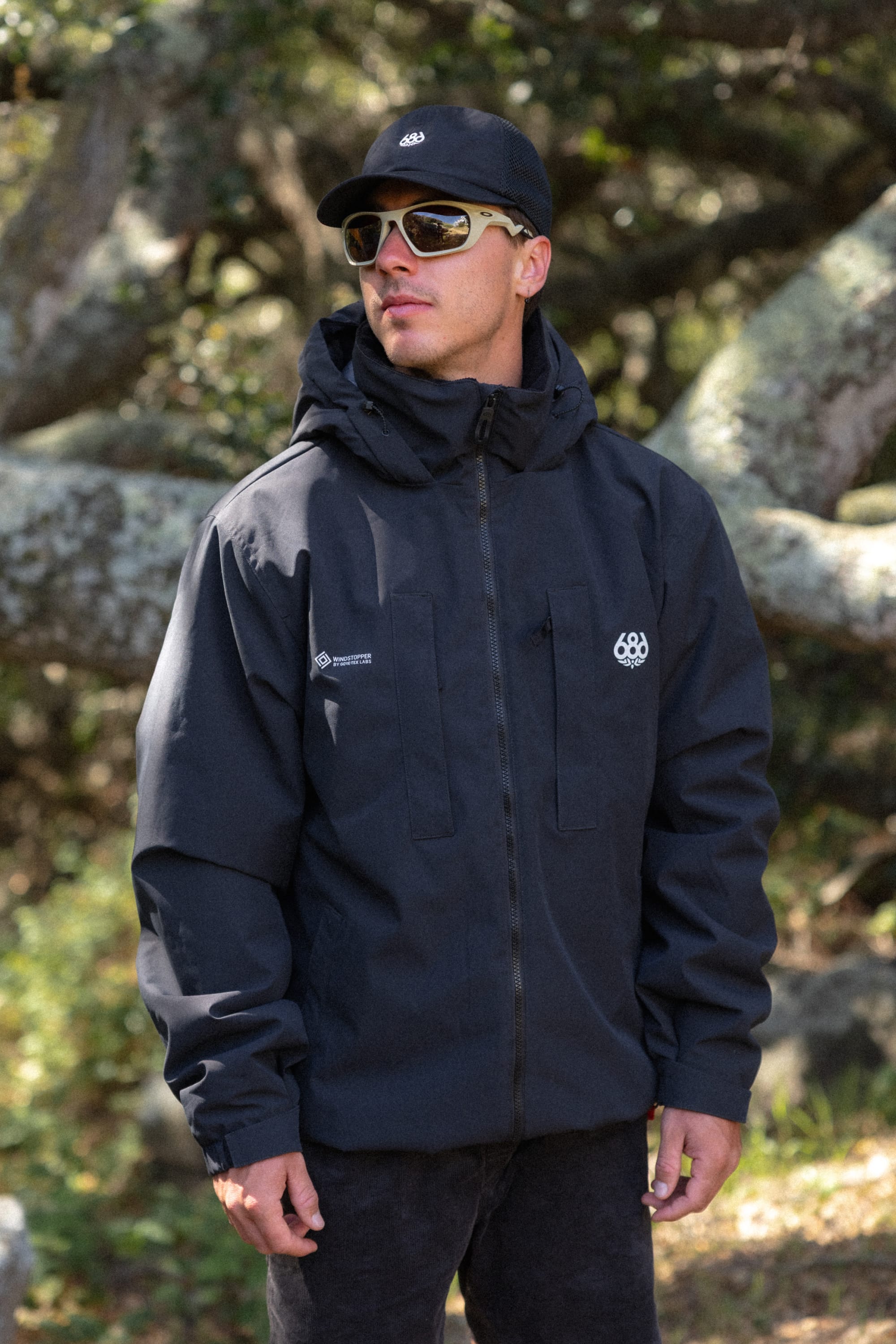 A man wearing the 686 Men’s WINDSTOPPER® Guideline Insulated Jacket by GORE-TEX® LABS stands outdoors in front of tree branches, paired with a black cap and sunglasses, looking to the side.