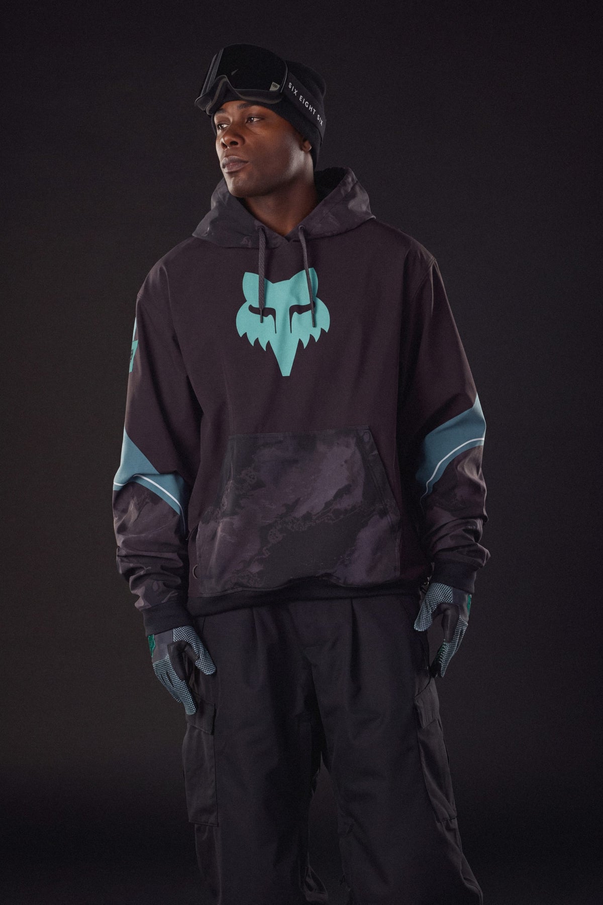 A man wearing a 686 Men’s Fox Racing Waterproof Team Hoody, a beanie, ski goggles, and gloves.