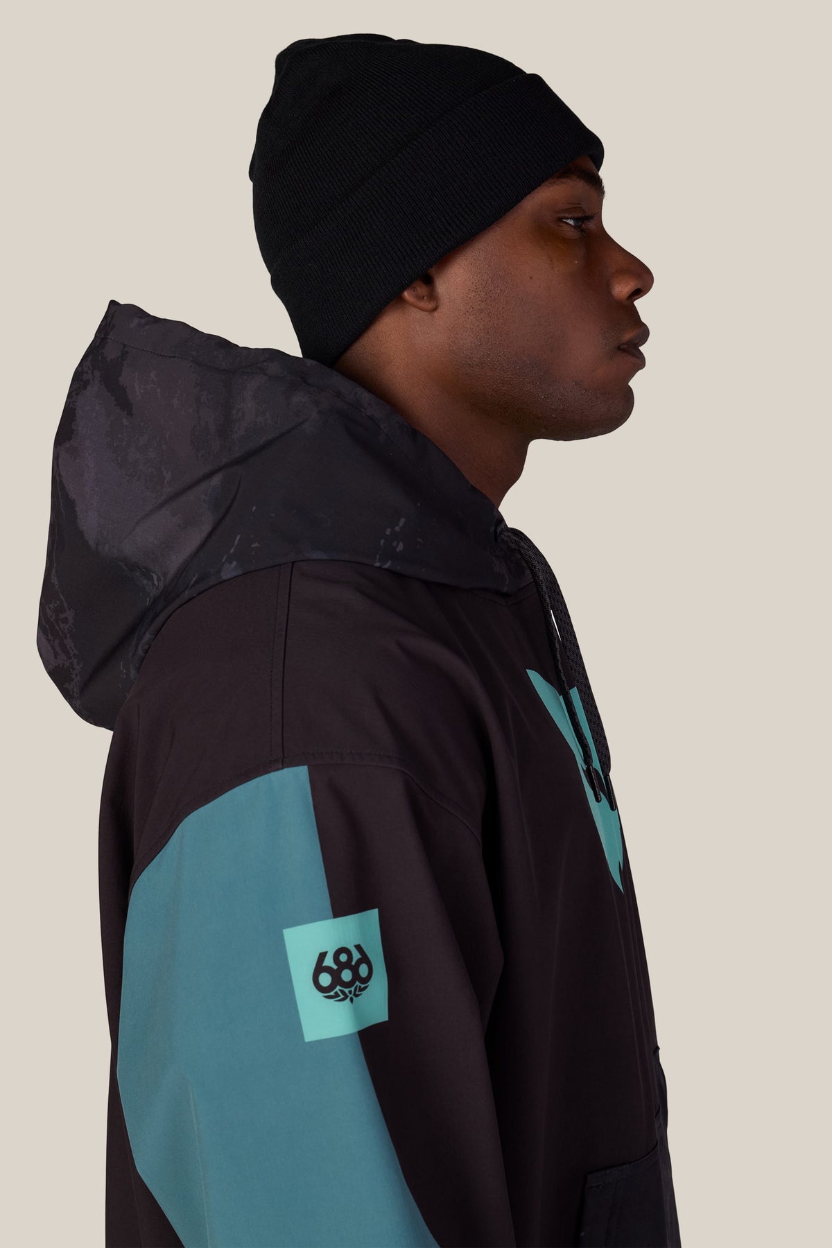 A man in profile wears a black beanie and a 686 Men’s Fox Racing Waterproof Team Hoody with teal accents and a 686 brand logo on the sleeve.