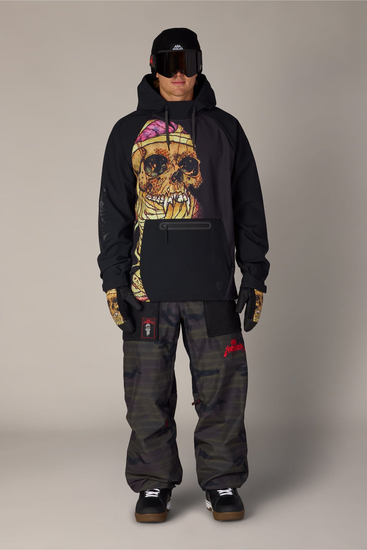 A person wearing a black 686 Men’s Metallica Waterproof Hoody with a large skull graphic on the chest, camouflage snow pants, ski goggles, and a beanie.