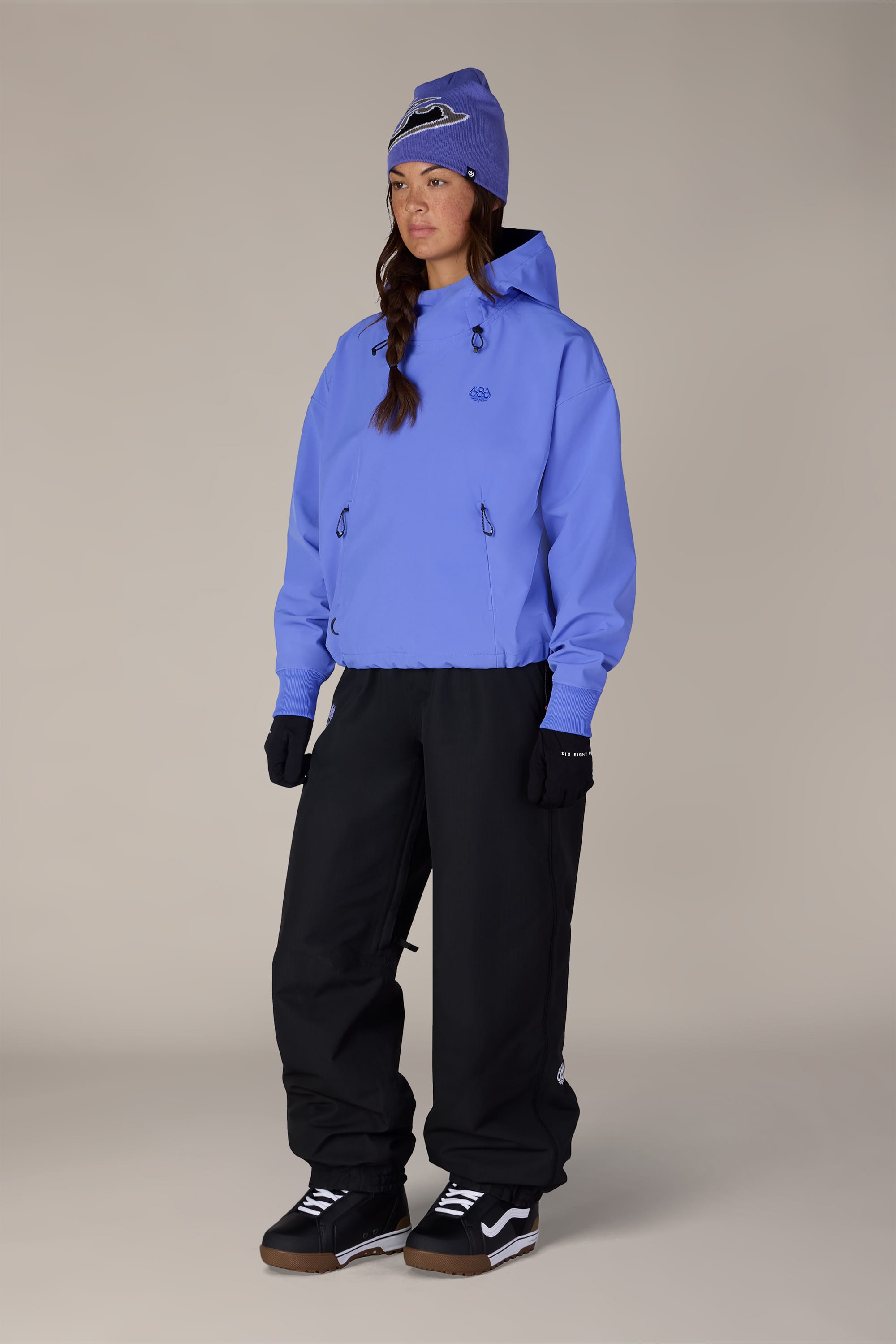 A person stands against a plain light background wearing a purple 686 Women’s Waterproof Team Crop Hoody, a matching beanie, black gloves, loose black pants, and black sneakers with white soles.