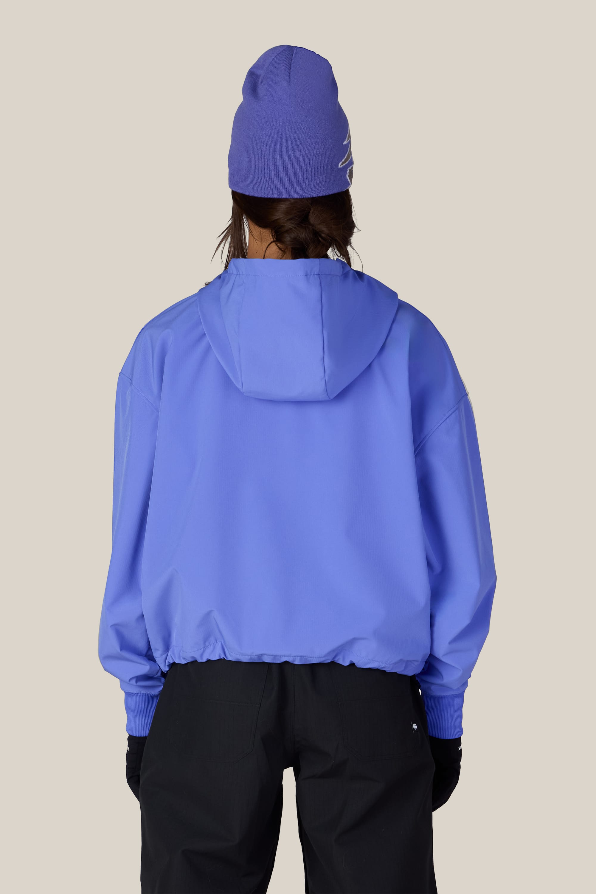 A person facing away from the camera is wearing the 686 Women’s Waterproof Team Crop Hoody in blue, paired with a matching blue beanie and black pants, set against a plain beige background.