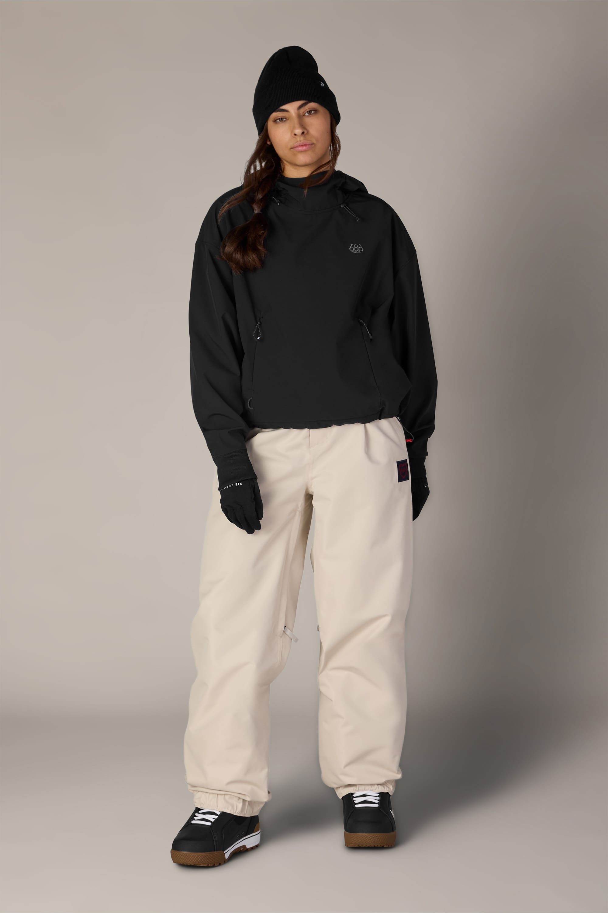 A person stands facing forward against a plain neutral background, wearing a black beanie, a black 686 Women’s Waterproof Team Crop Hoody layered under a black jacket, black gloves, beige baggy pants, and black sneakers with white soles.