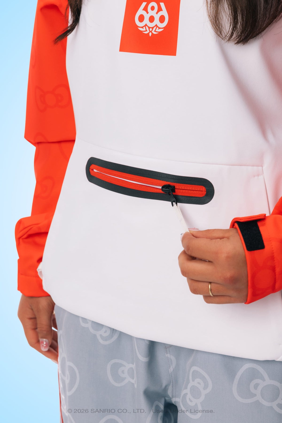 A person wearing the 686 Women’s Hello Kitty® Waterproof Hoody, featuring a white and orange woven design with a red zipper pocket, is pulling the zipper. Their hand, adorned with a gold ring, is visible. They are also wearing light blue-gray pants with a subtle pattern.