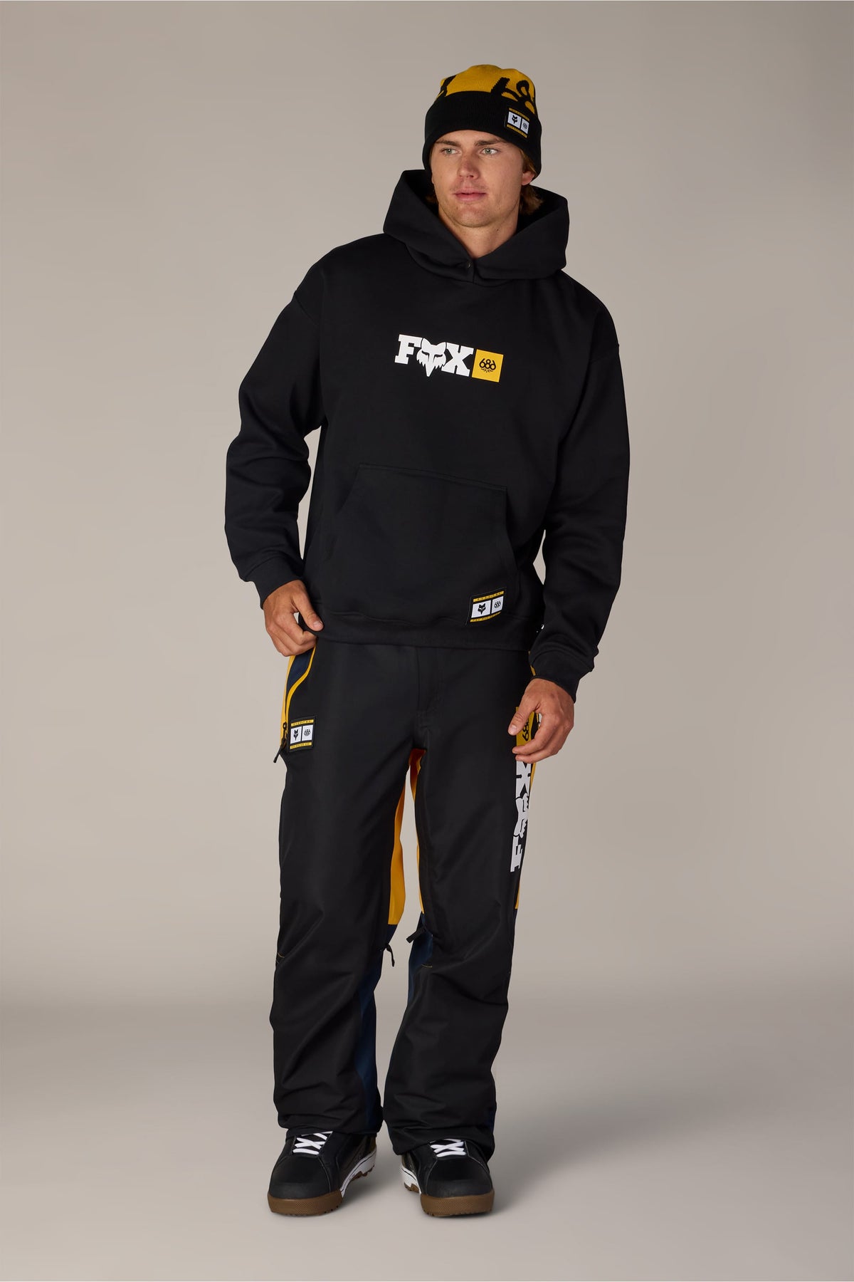A man wearing a 686 Men’s Fox Racing Premium Heavyweight Pullover Hoody, matching snow pants with yellow accents, and a branded beanie.