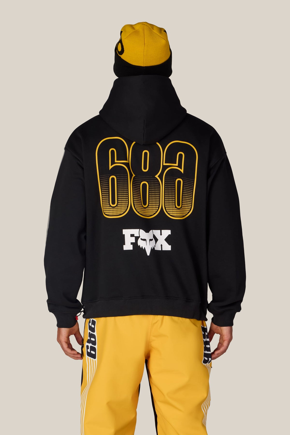 A person seen from behind wearing a 686 Men’s Fox Racing Premium Heavyweight Pullover Hoody paired with yellow pants featuring black and white side stripes.