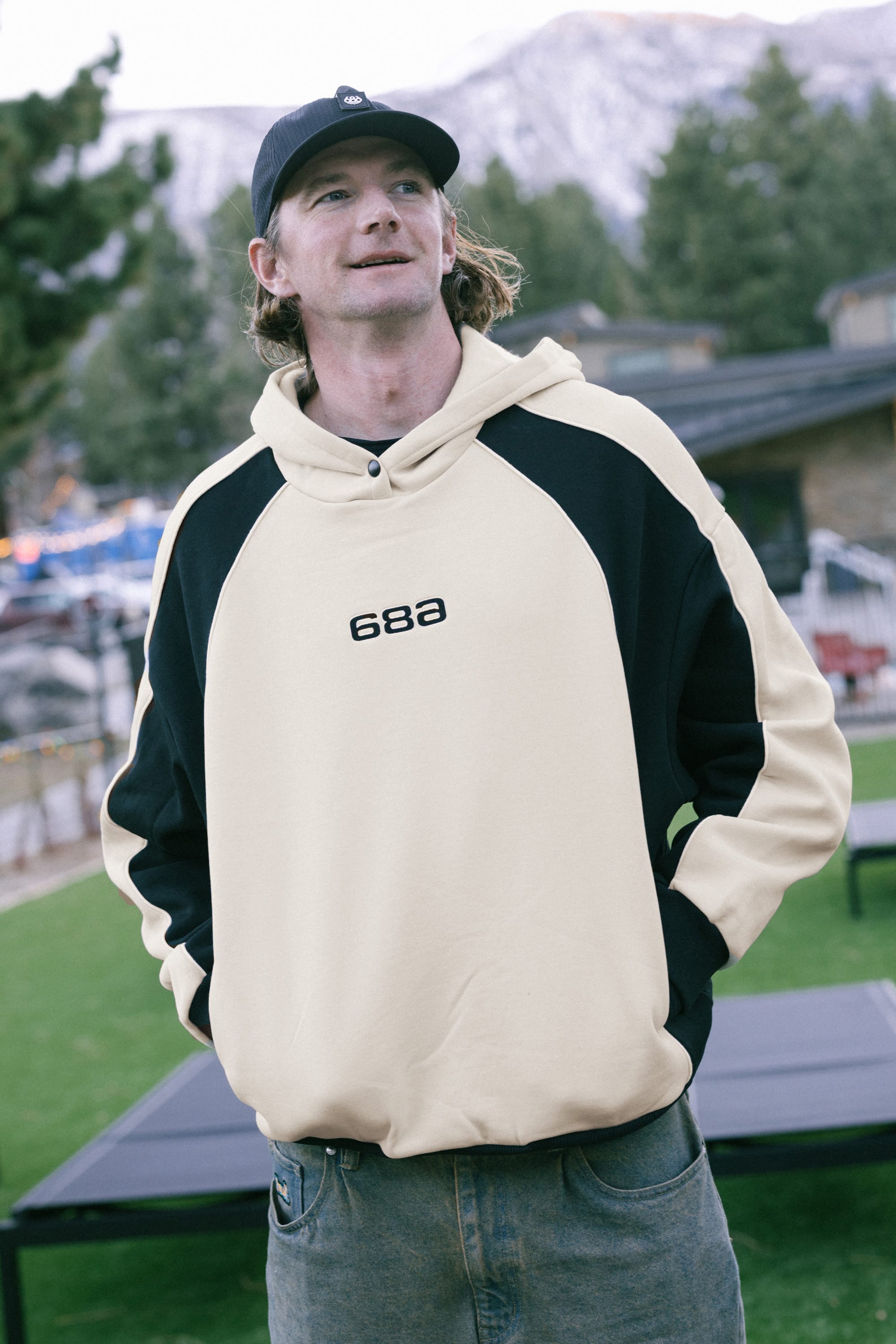 Wearing the 686 Legacy Premium Heavyweight Hoody, a man stands outdoors on green grass with mountains, trees, and buildings in the background, smiling slightly and looking upward.