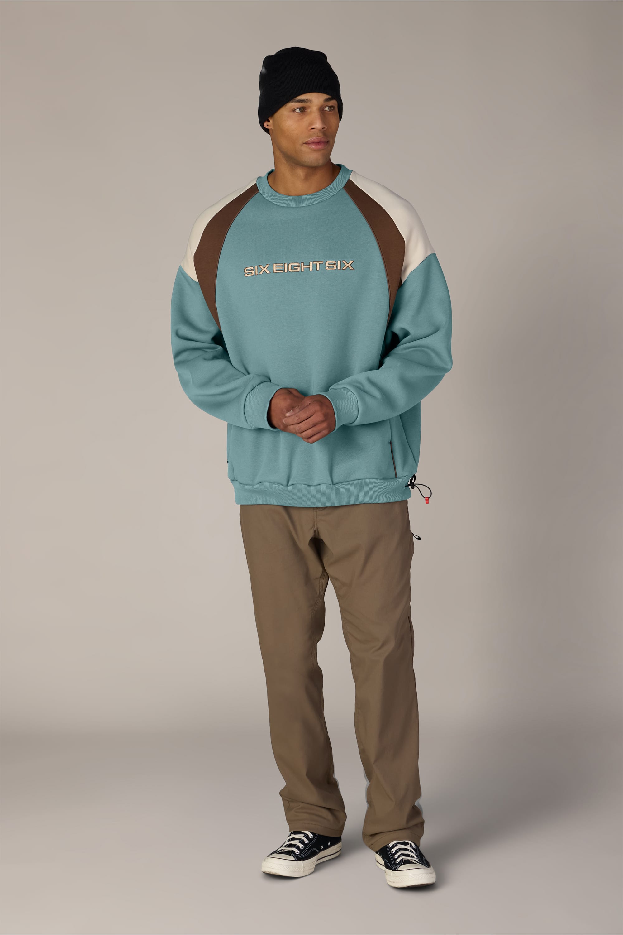 Wearing the 686 Reserved® Premium Heavyweight Crewneck by 686—a teal and brown men’s sweatshirt crafted from soft cotton—a person stands against a plain background, paired with a black beanie, tan pants, and black-and-white sneakers. They look slightly to the side with their hands clasped.