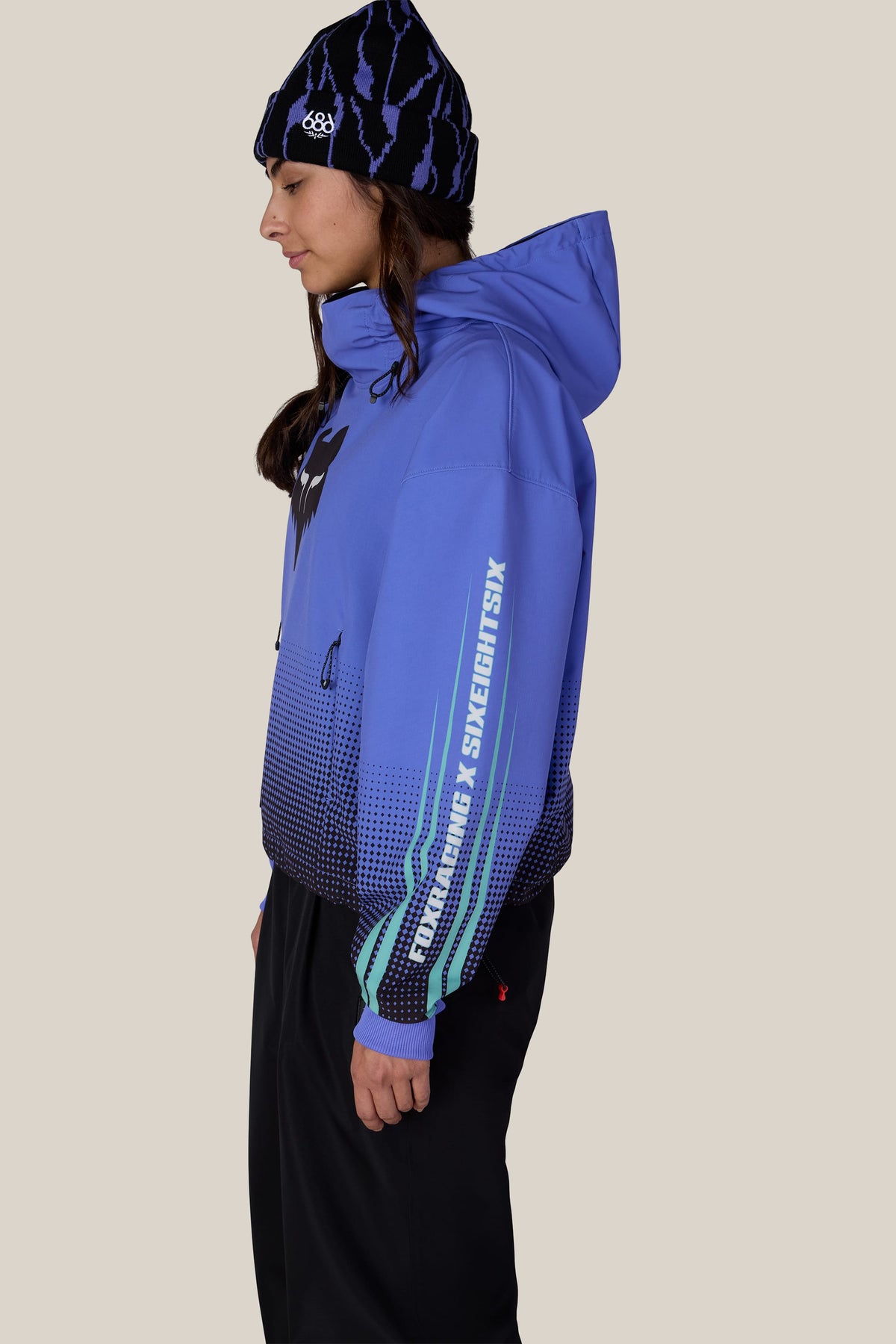 A person in profile wears a blue 686 beanie and a blue 686 Women’s Fox Racing Waterproof Crop Hoody featuring a black gradient pattern and logo text down the sleeve.
