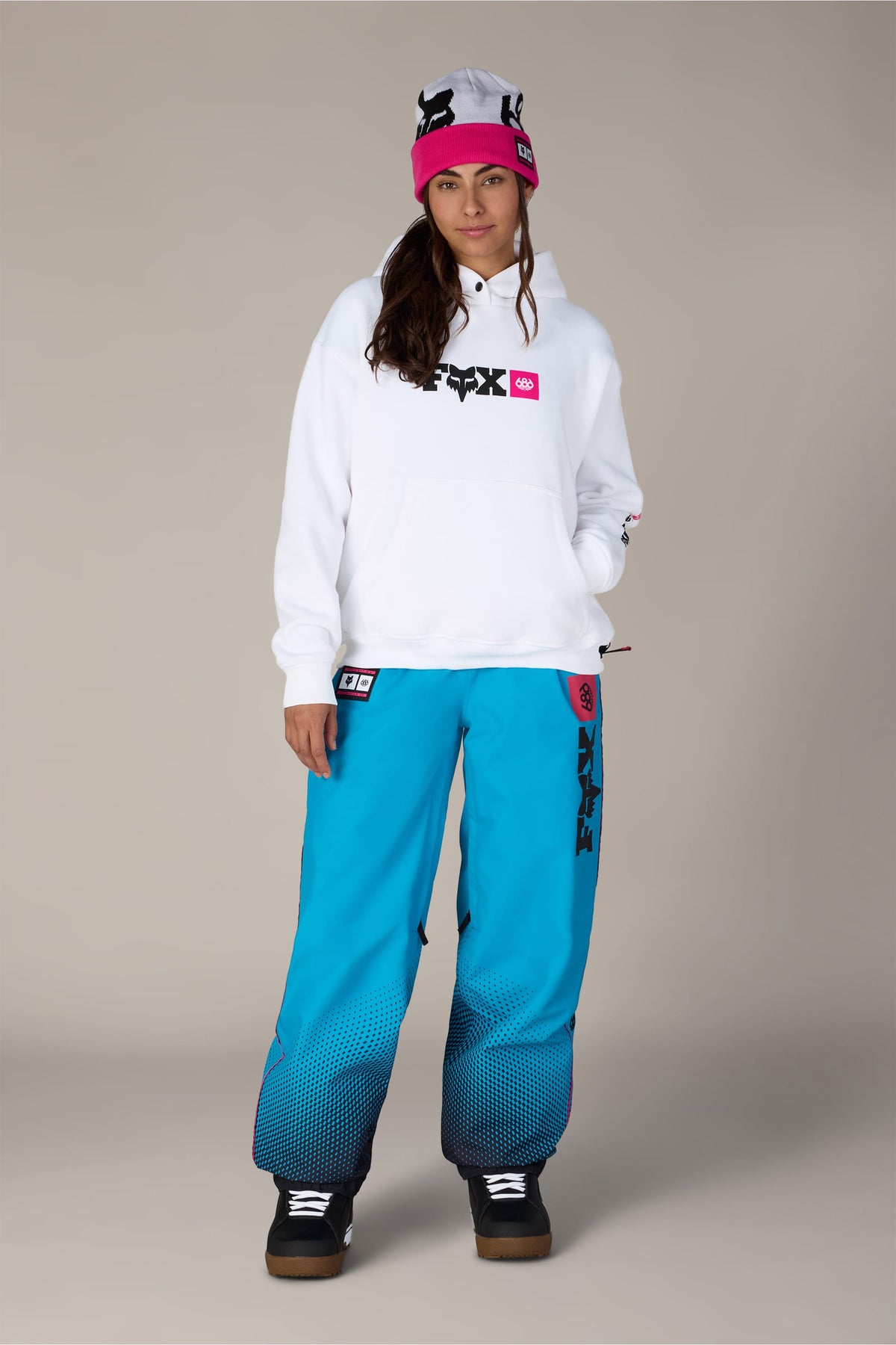 A woman wearing a pink beanie, a 686 Women’s Fox Racing Premium Heavyweight Pullover Hoody, and blue snow pants with a black gradient pattern at the cuffs.