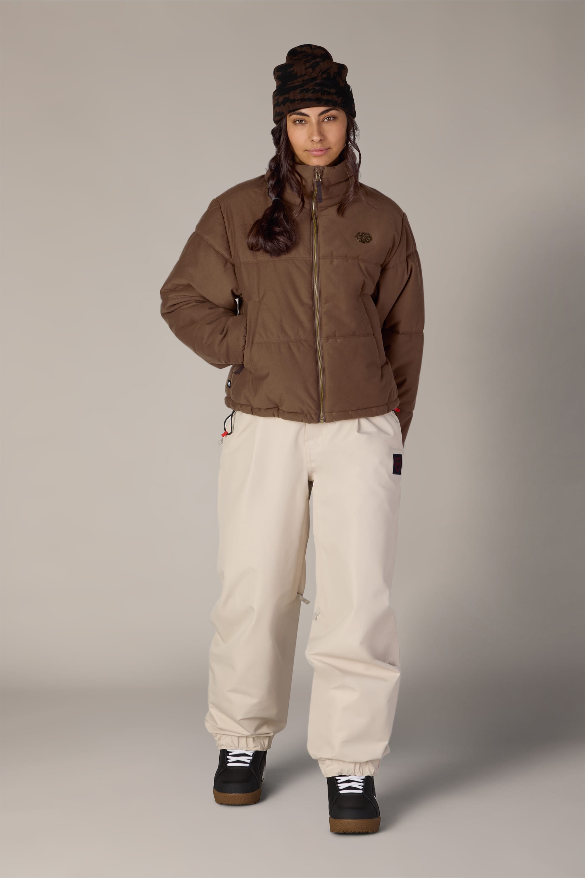 Wearing the 686 Women’s Gracious Puffer Jacket by 686, along with cream baggy pants, black boots, and a brown beanie, a person stands against a plain light gray background, looking at the camera with one hand in their pocket.